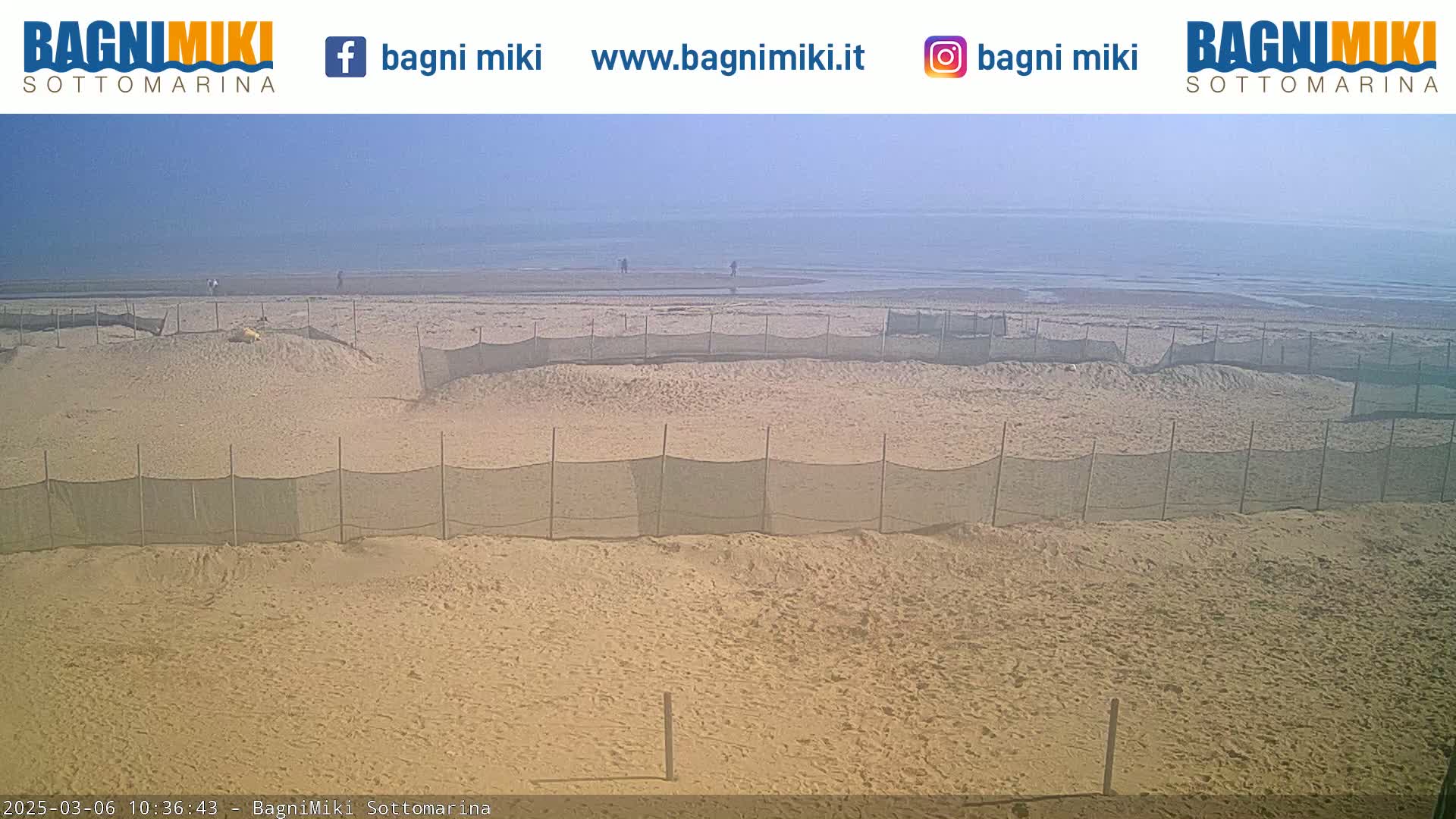 A sandy beach with several fenced-off areas stretches to a calm, hazy ocean under a clear sky.