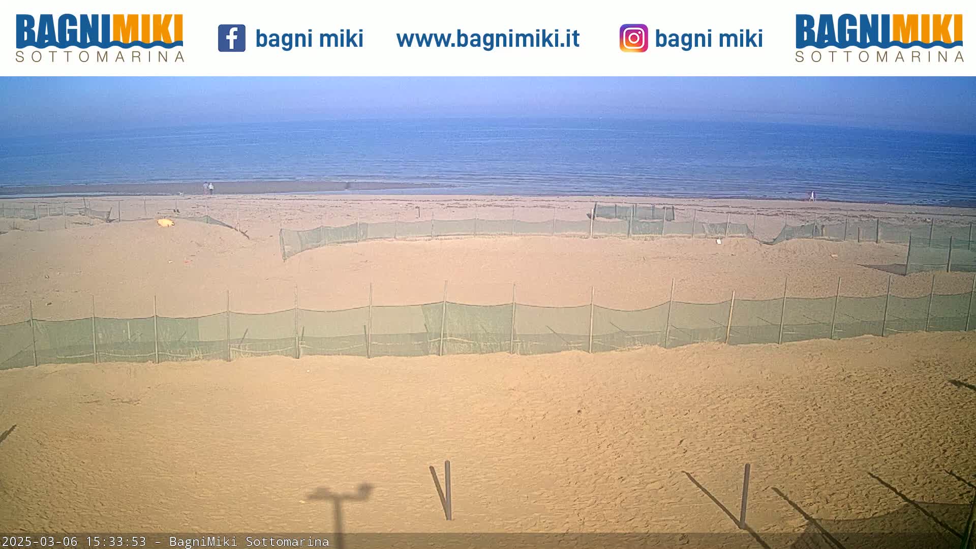 A sandy beach with several enclosed areas, protected by netting, stretches along a calm, blue ocean under a clear sky.