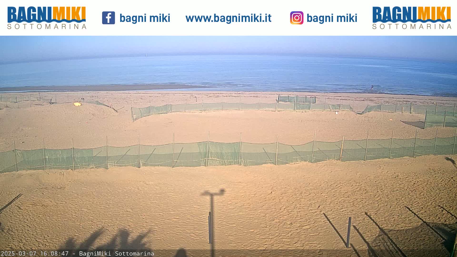 A sandy beach with several fenced-off areas is visible, under a mostly clear sky, with calm ocean water in the background.