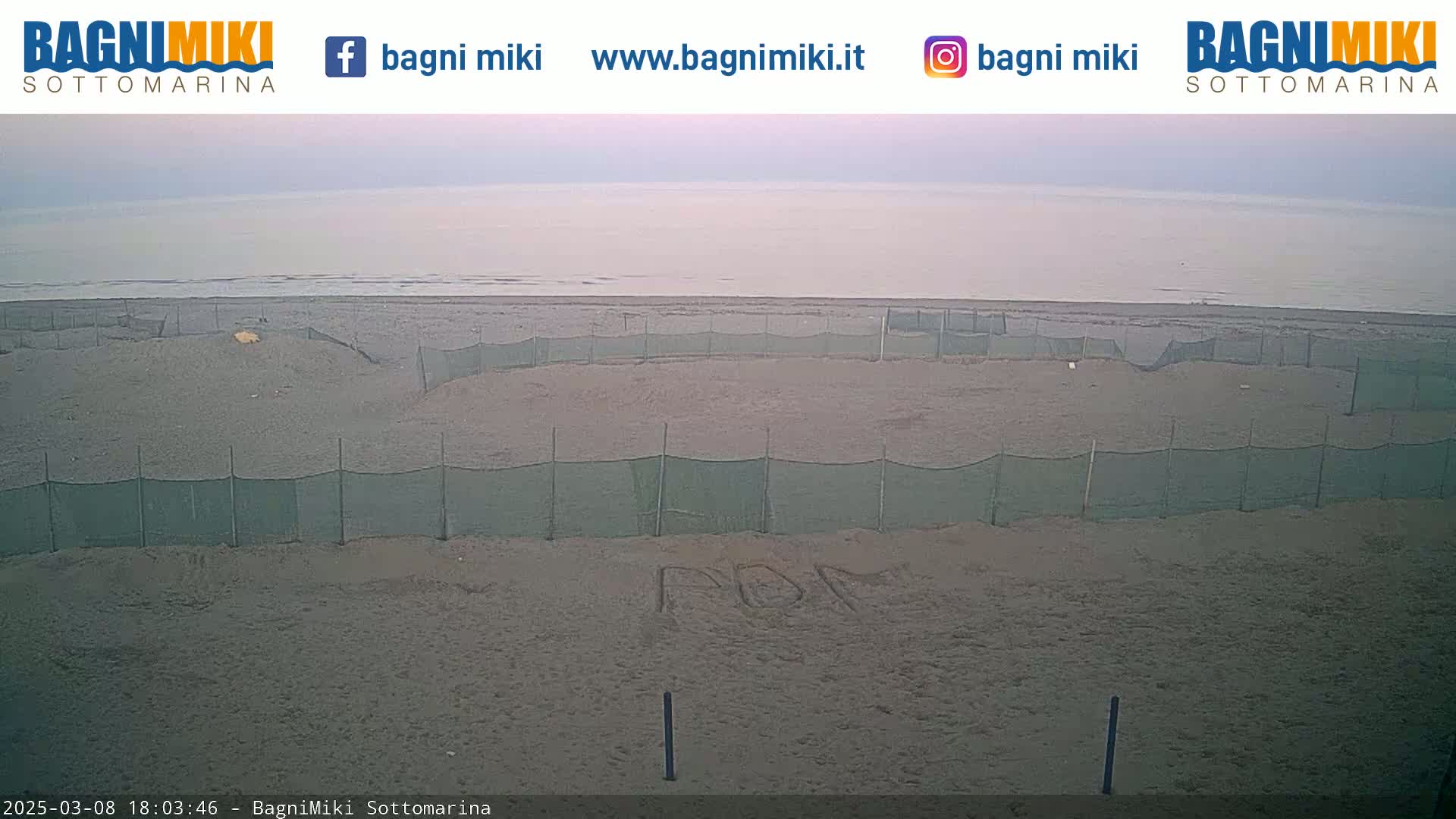 A sandy beach with green netting sections and the faint lettering "ROM" written in the sand is visible under a mostly cloudy sky; a calm sea is in the background.