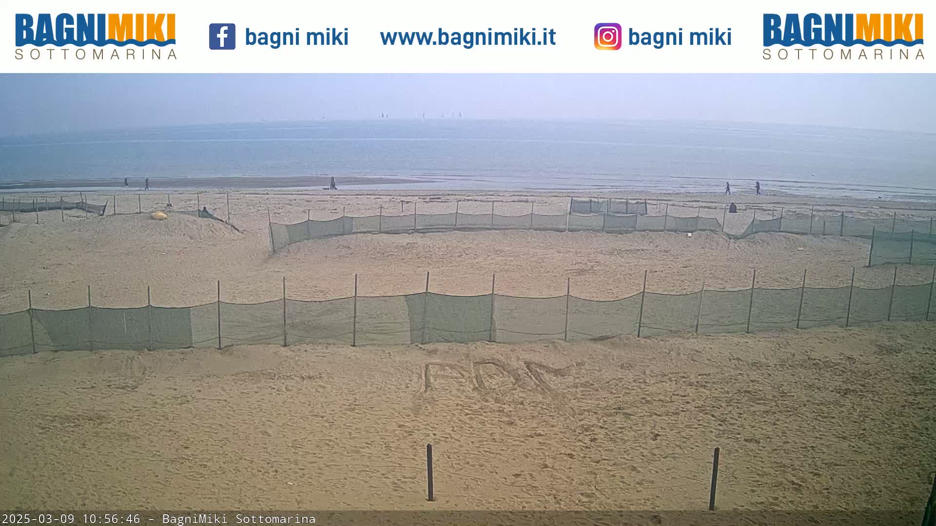 A sandy beach with several netted enclosures and a hazy, calm sea in the background.