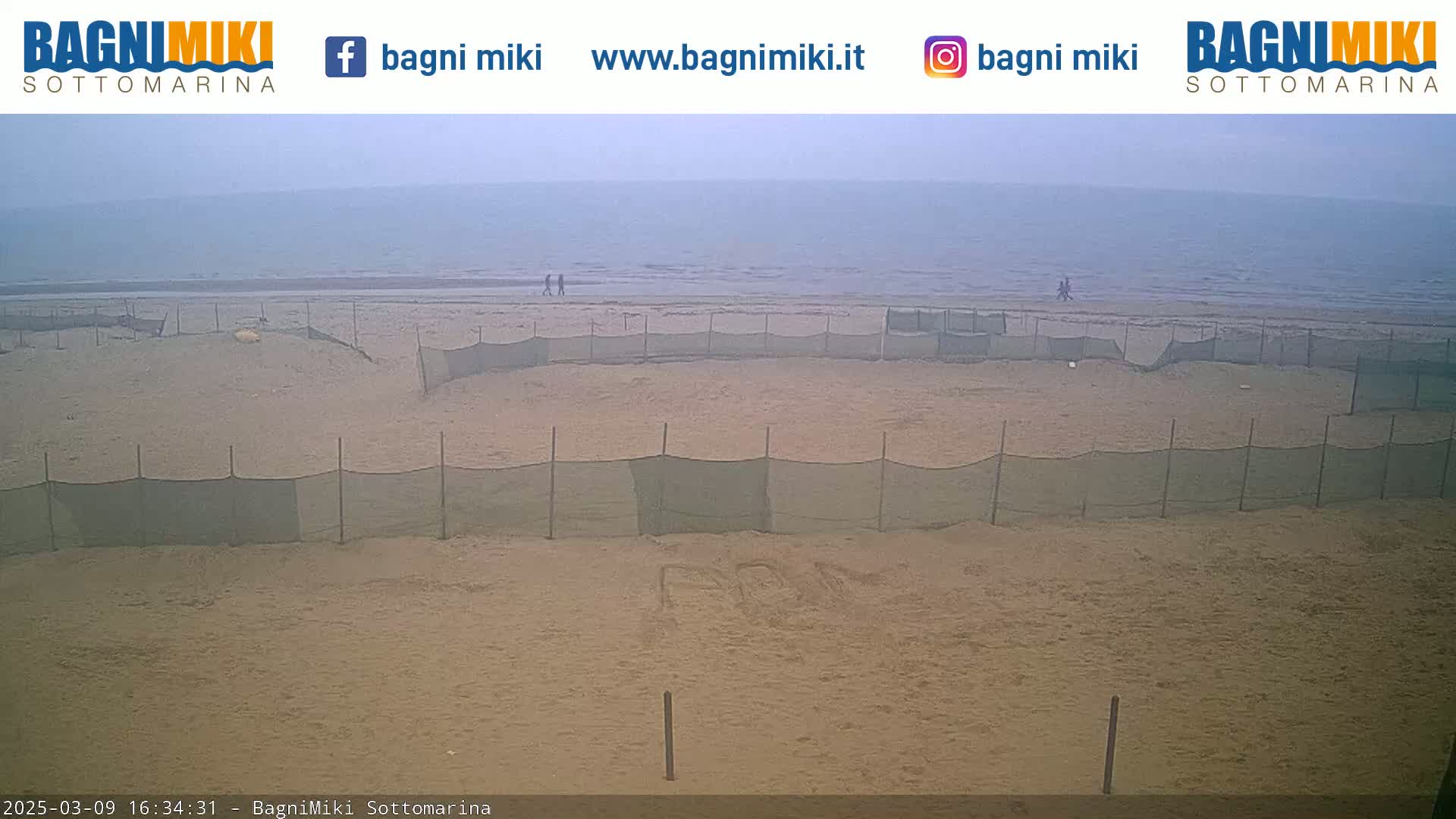 A sandy beach with several sections enclosed by netting is visible, overlooking a calm, hazy ocean.