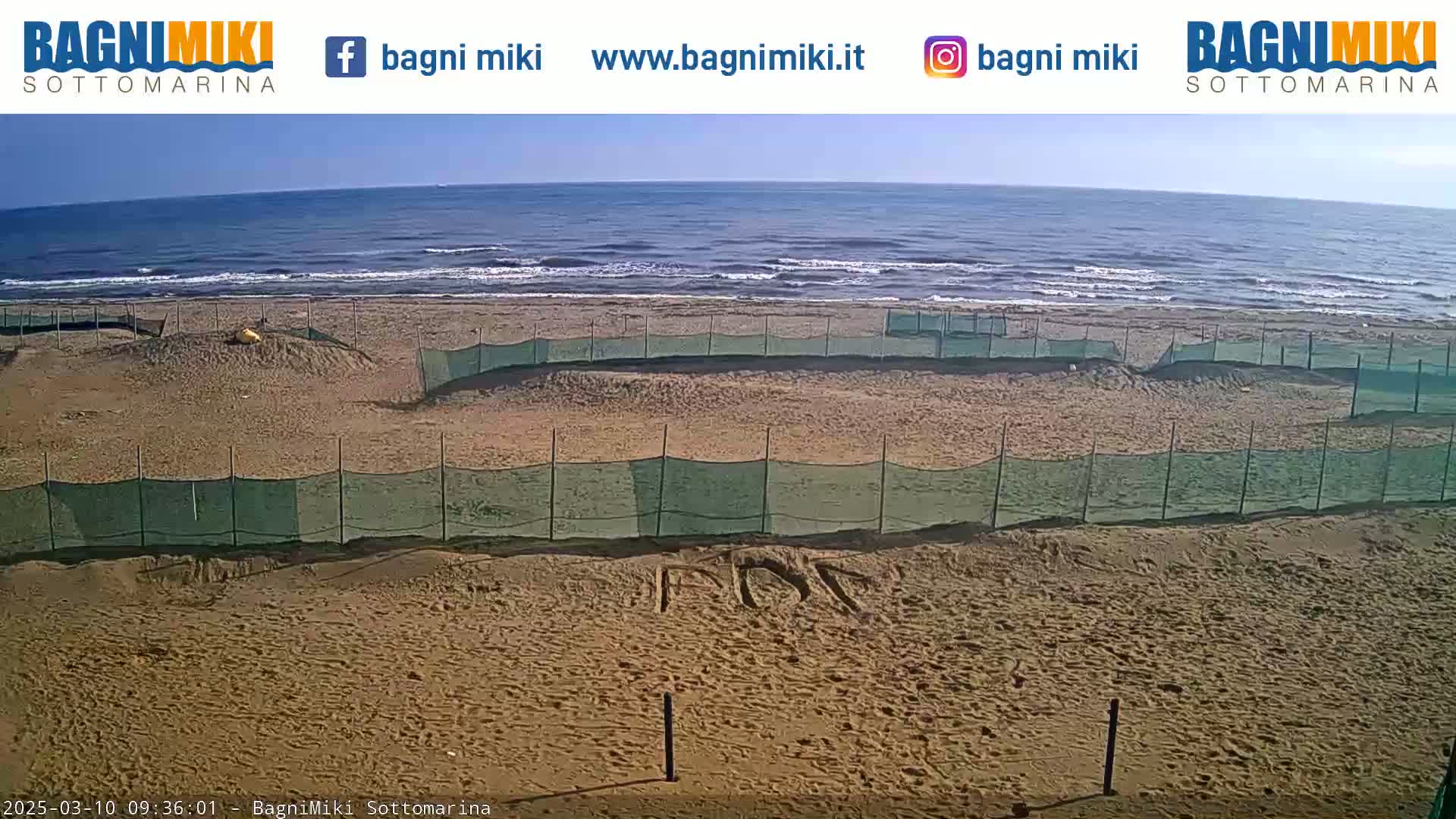 A sandy beach with several enclosed areas marked by green netting is visible, overlooking a calm ocean under a sunny sky.