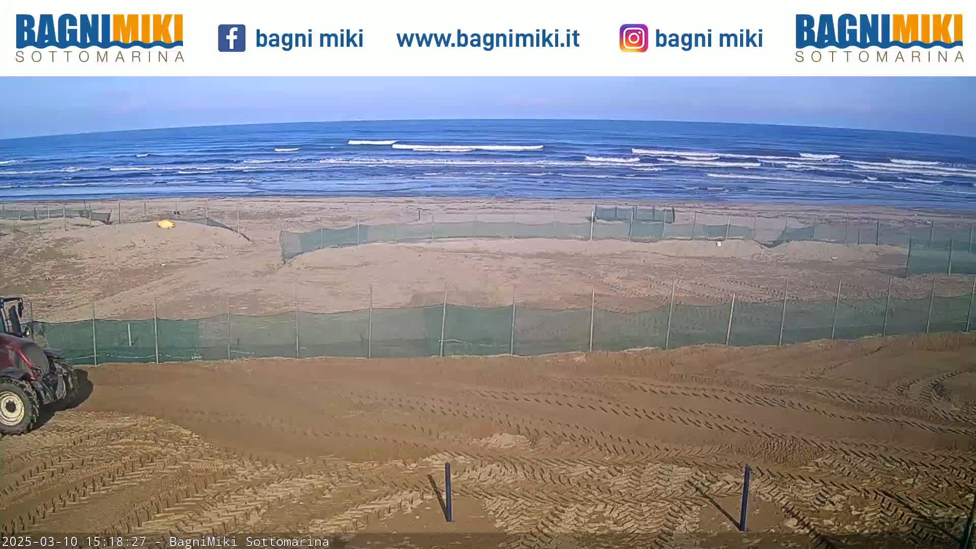 A tractor is parked near a sandy beach with calm ocean waves and several fenced-off areas on the sand.