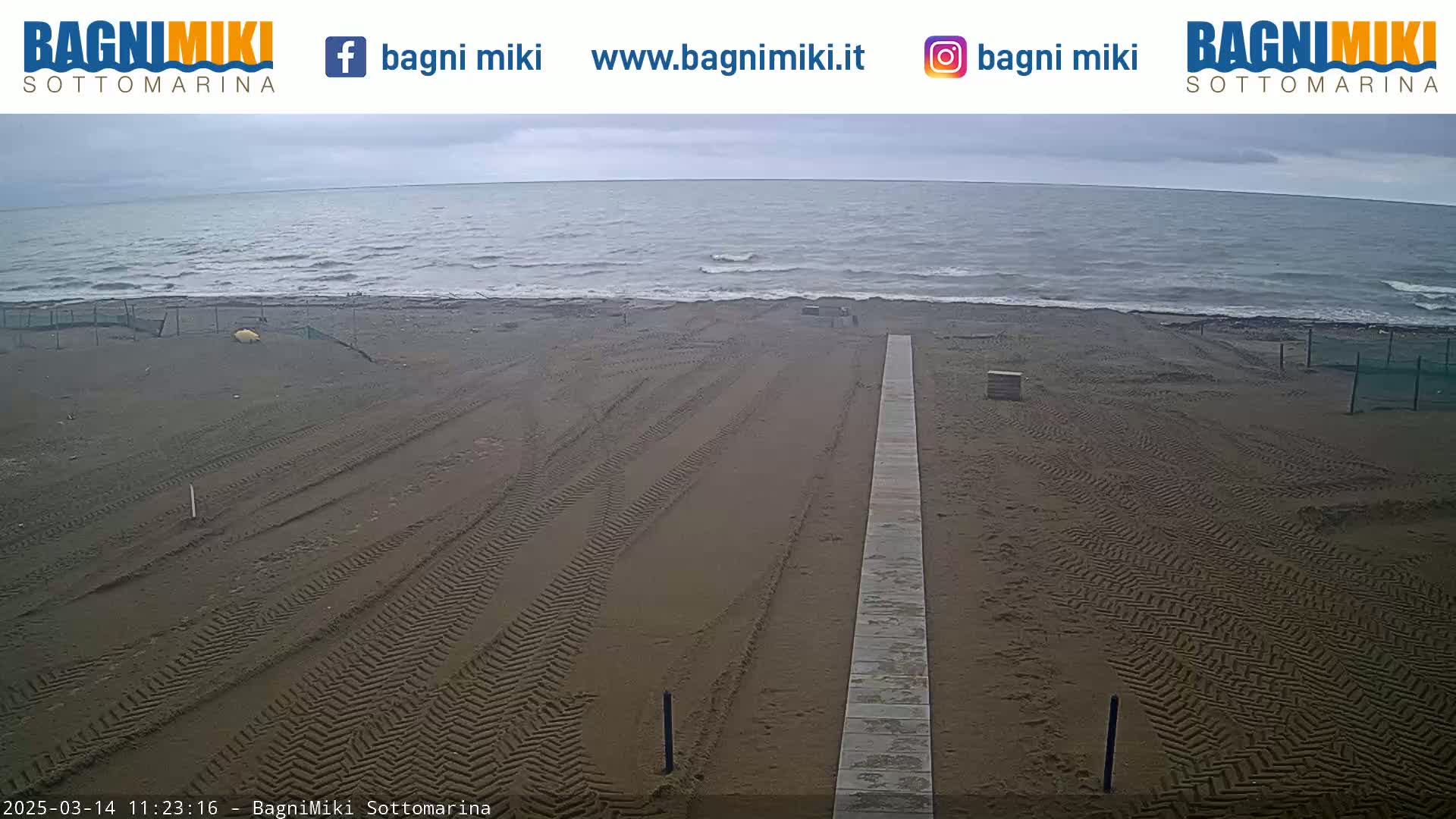 A sandy beach with tire tracks and a long boardwalk stretches toward a gray, wavy sea under an overcast sky.