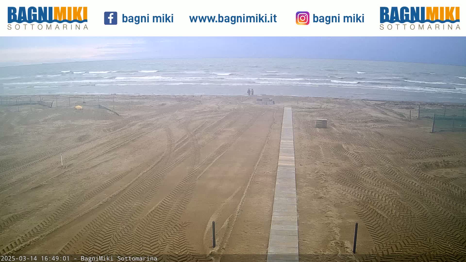 A sandy beach with tire tracks leading to a boardwalk stretches towards a wavy ocean under an overcast sky.