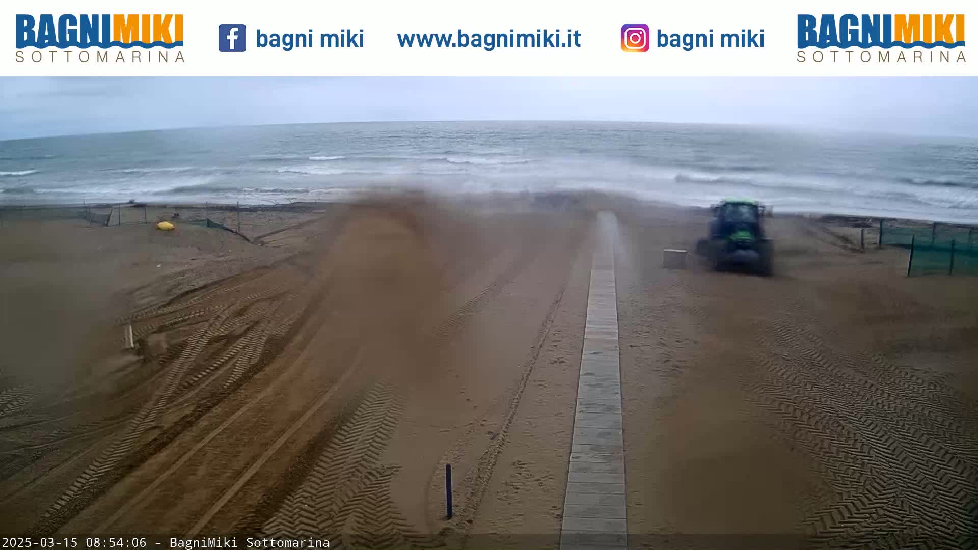 A tractor is driving on a sandy beach near a wavy, overcast ocean.