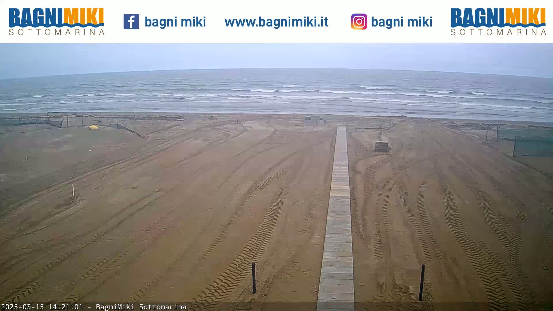 A sandy beach with tire tracks leading to a wooden walkway, under an overcast sky, overlooks a wavy ocean.