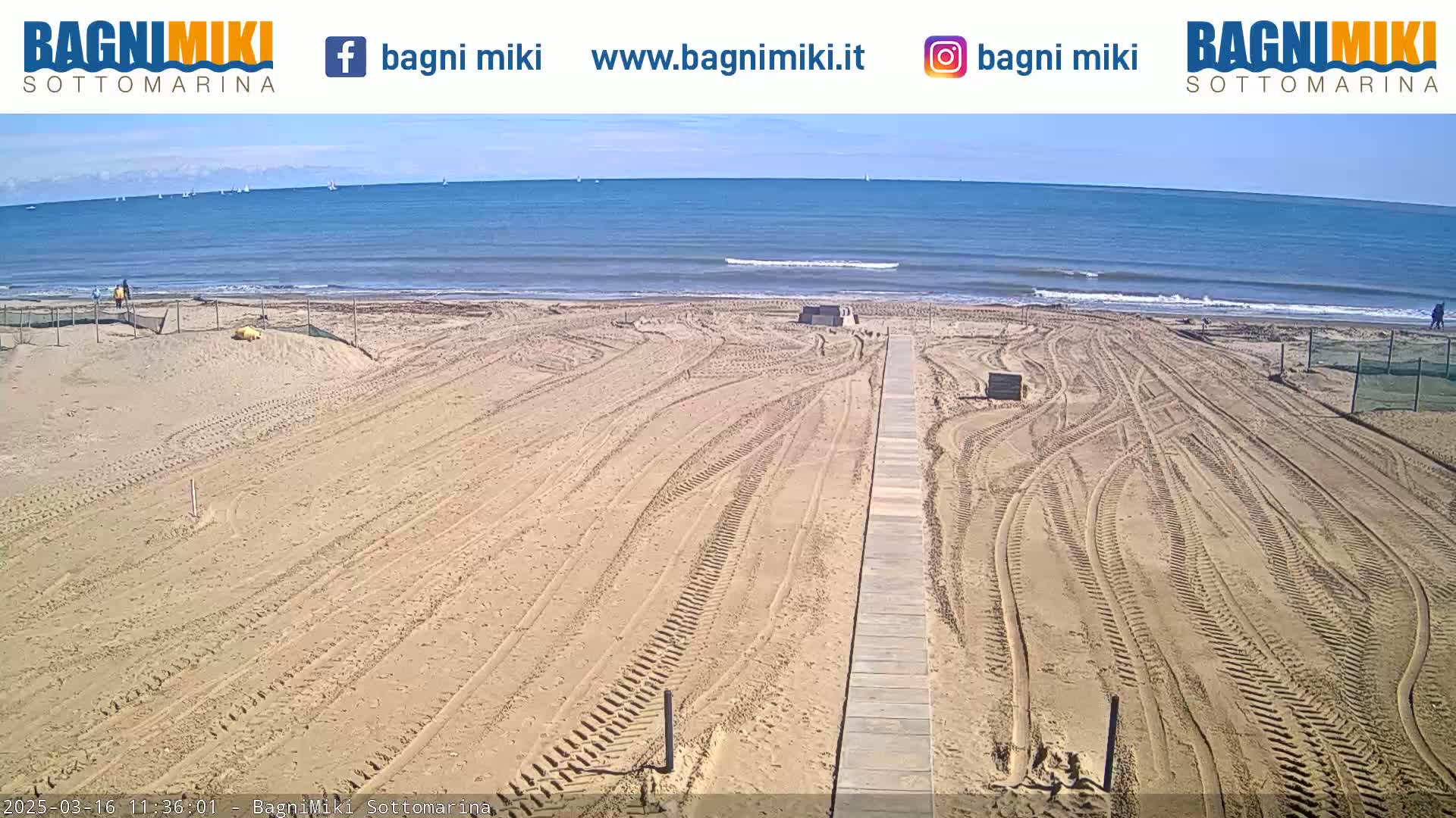 A sandy beach with tire tracks leading to a wooden boardwalk, under a clear sky and calm ocean in the background.