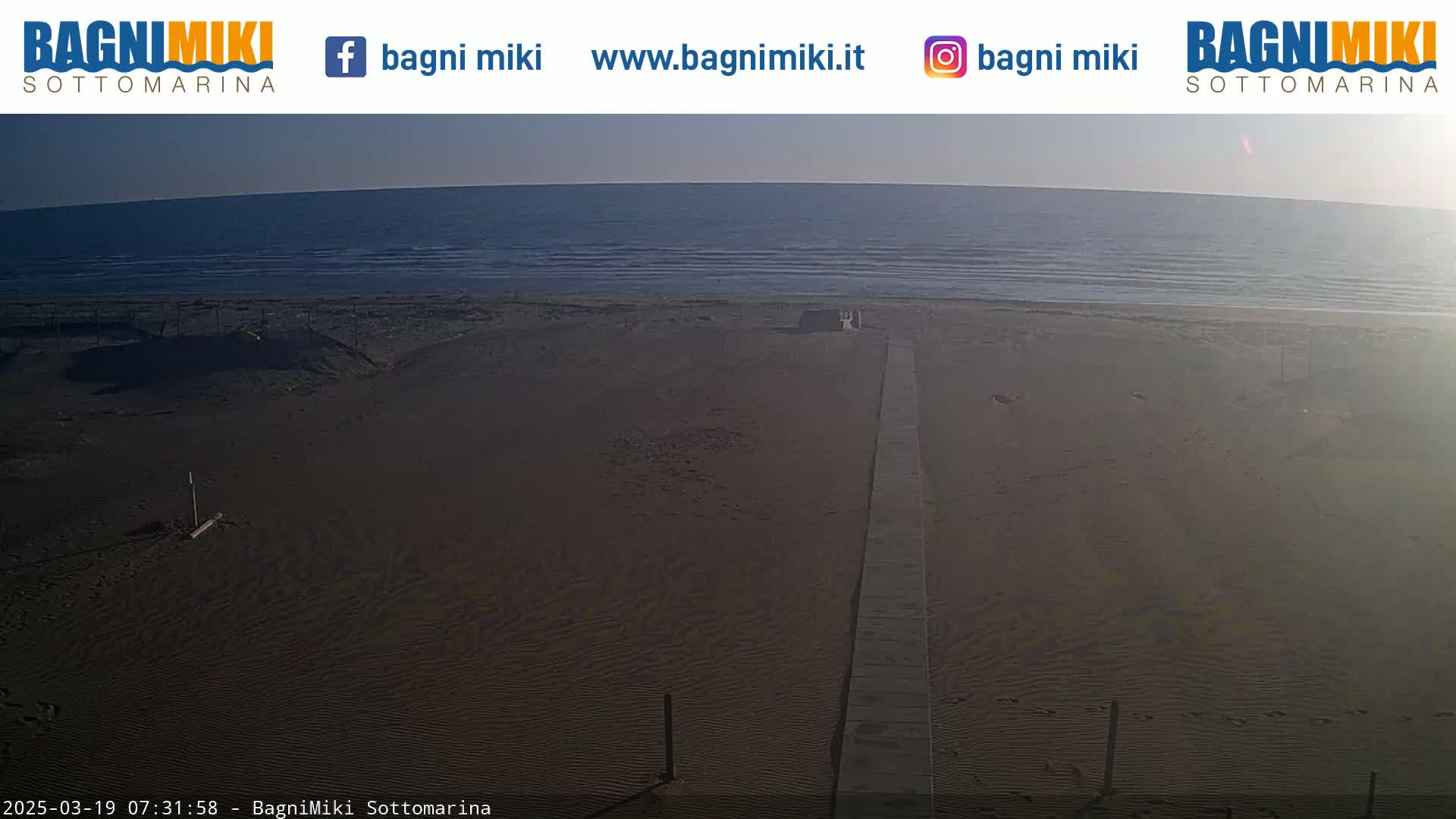 A long wooden walkway leads from a sandy beach to a calm ocean under a clear sky.