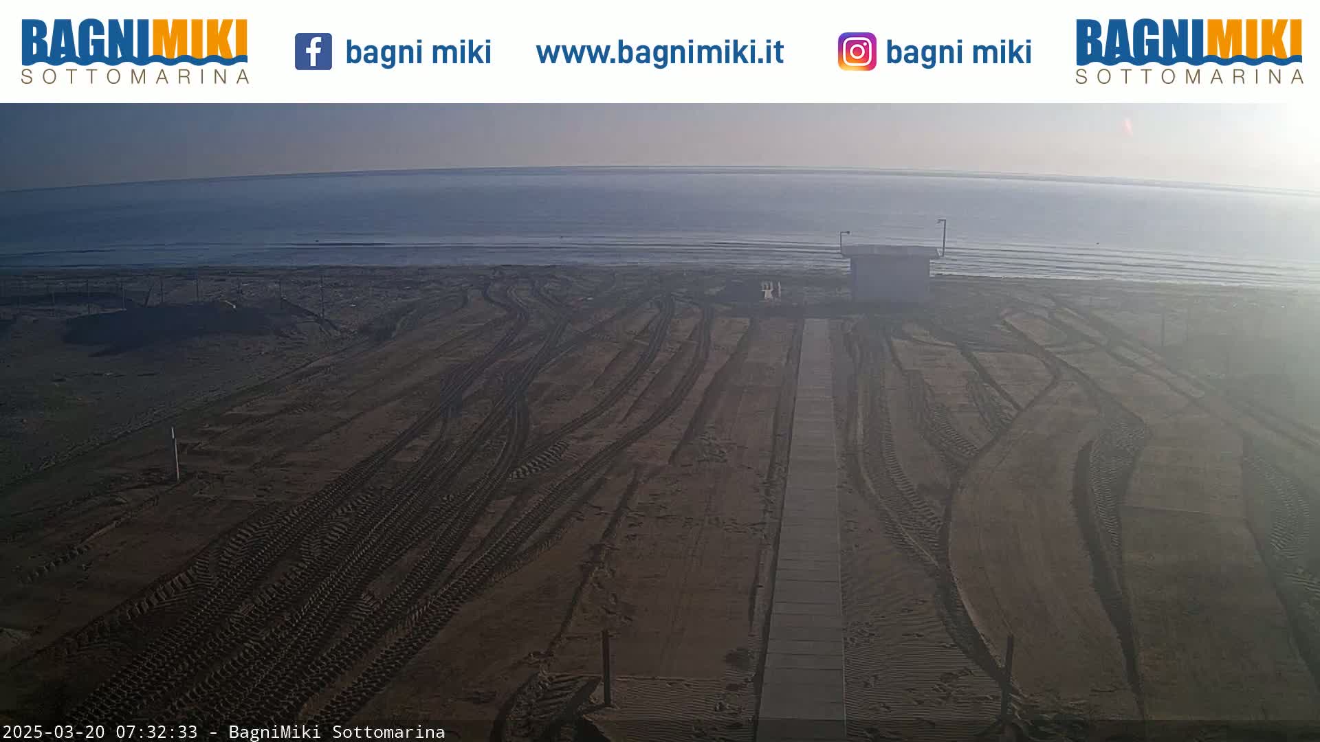 A sandy beach with tire tracks leading to a small building next to a calm ocean under a hazy sky.