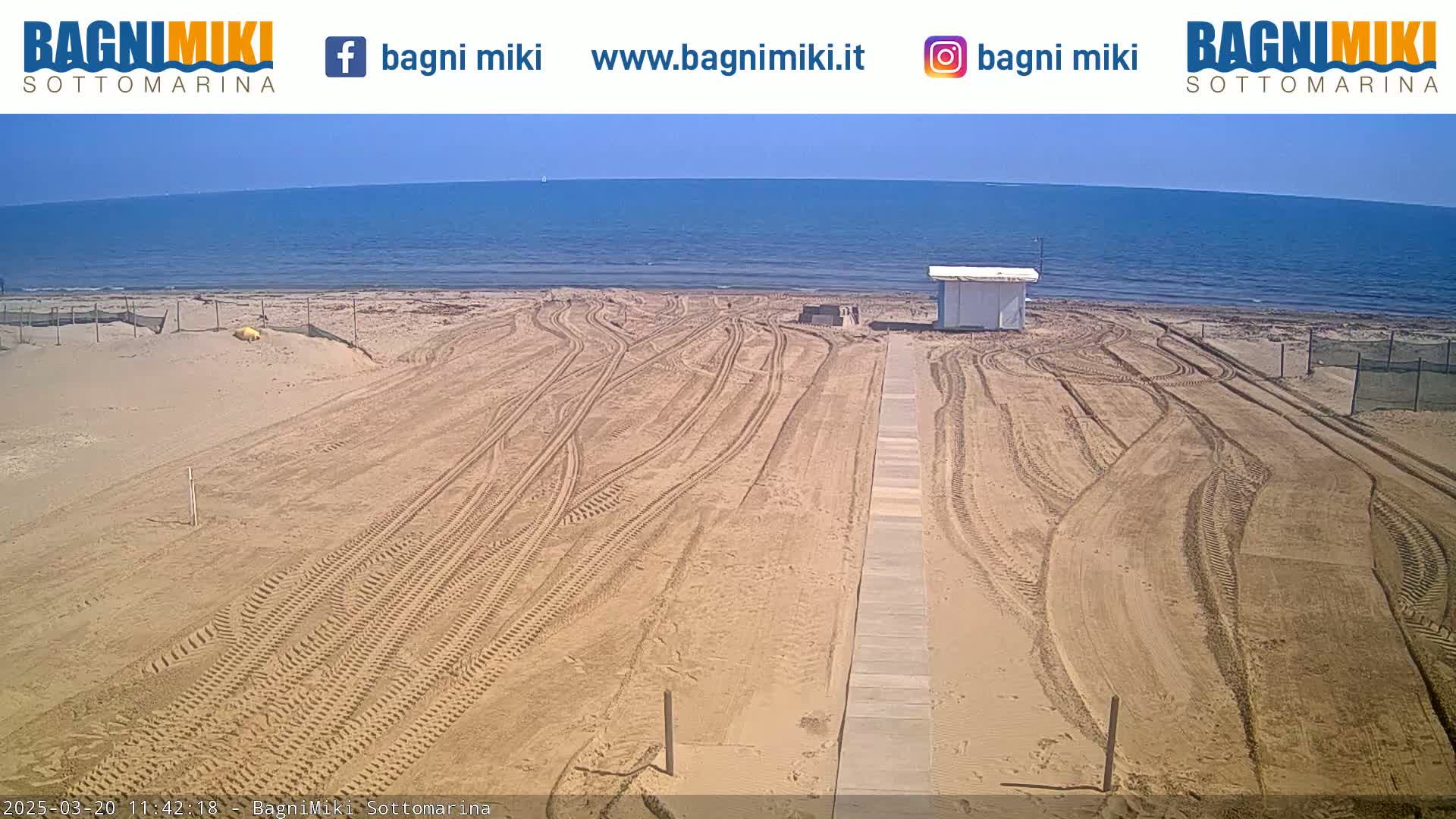 A sandy beach with tire tracks leading to a small building near a calm, blue ocean under a clear sky.
