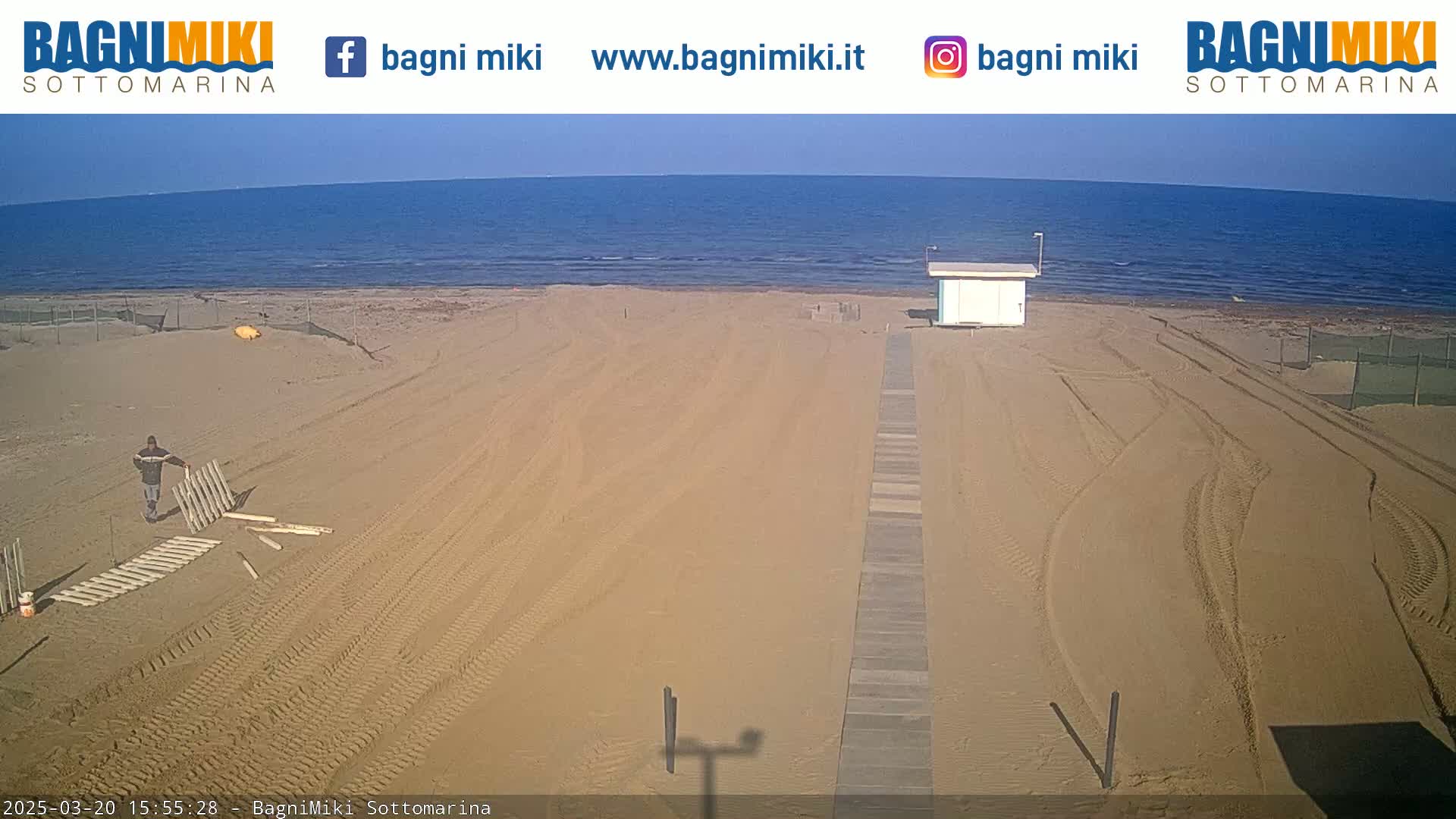 A sandy beach with tire tracks and a person assembling a wooden fence is visible under a sunny sky next to a calm ocean.