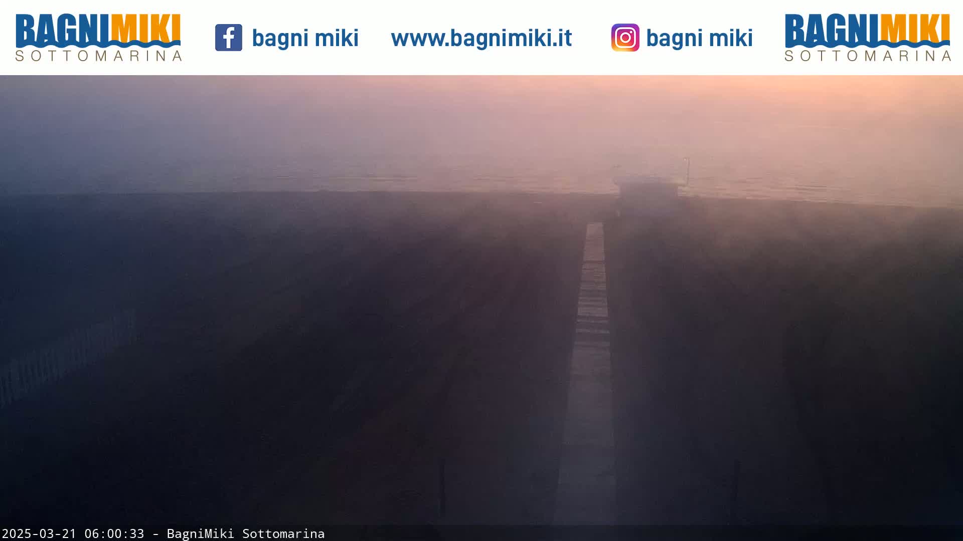 A hazy sunrise illuminates a long, narrow walkway leading to a body of water under a misty sky.