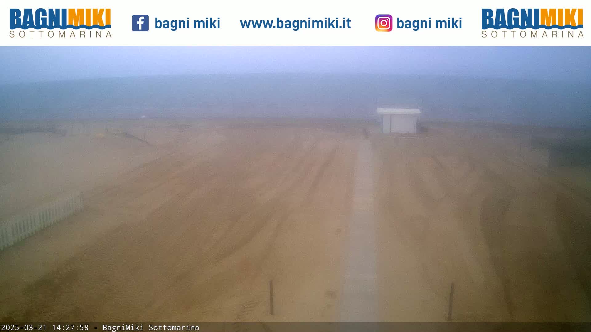 A hazy, overcast day reveals a sandy beach with tire tracks leading to a small structure near the ocean.