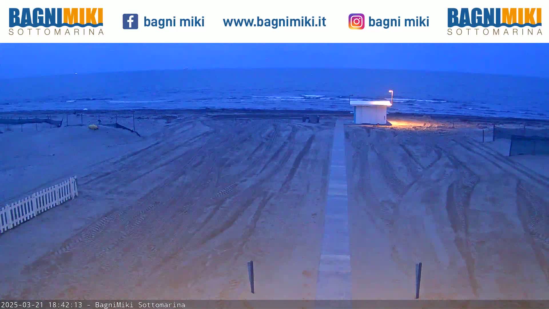 A mostly empty sandy beach at dusk is shown with tire tracks leading to a small structure near the water's edge.