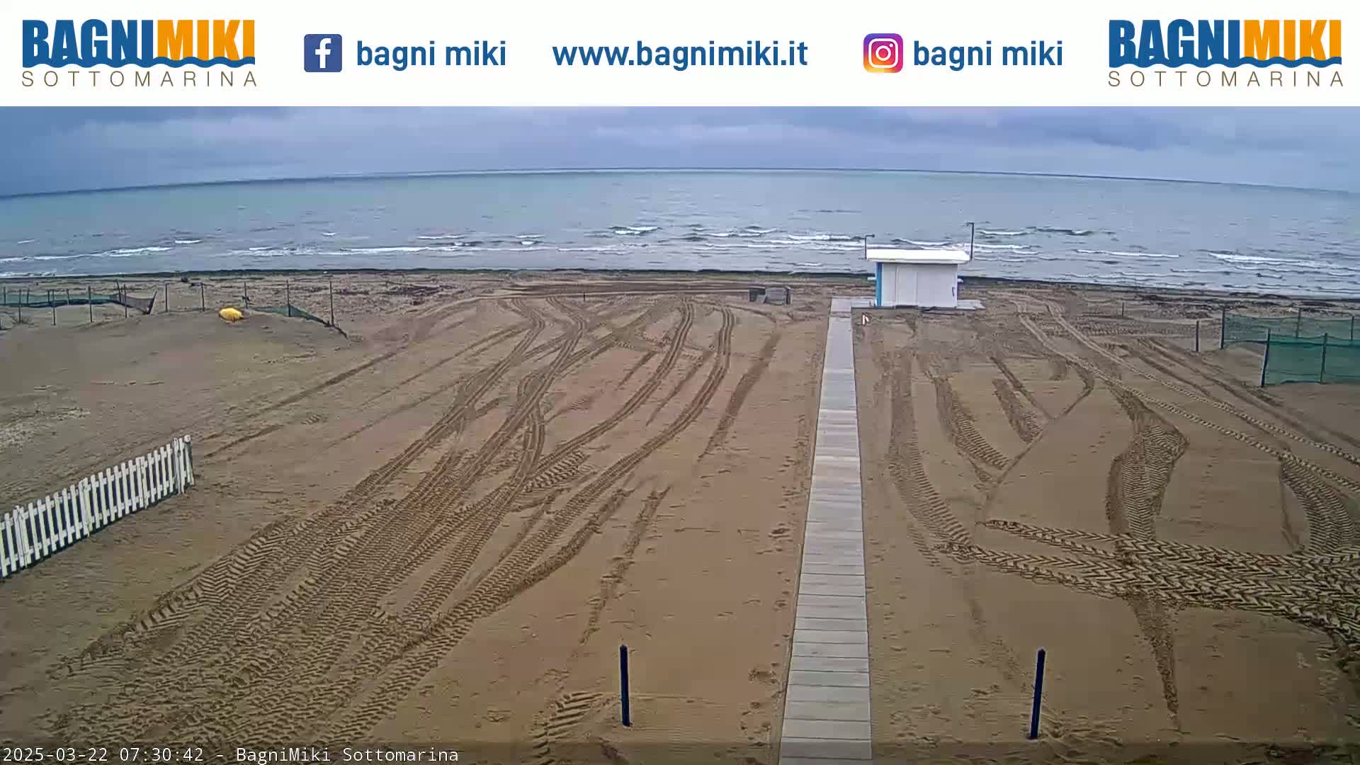 A mostly cloudy day at a sandy beach shows tire tracks crisscrossing the sand leading to a boardwalk toward a small building near the ocean.