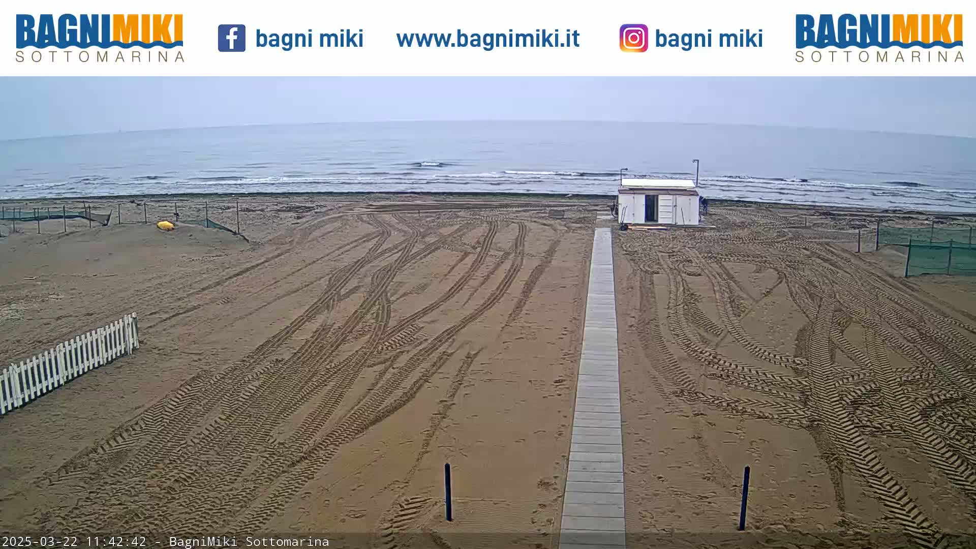 A sandy beach with tire tracks leading to a small building is visible under an overcast sky, next to a calm ocean.