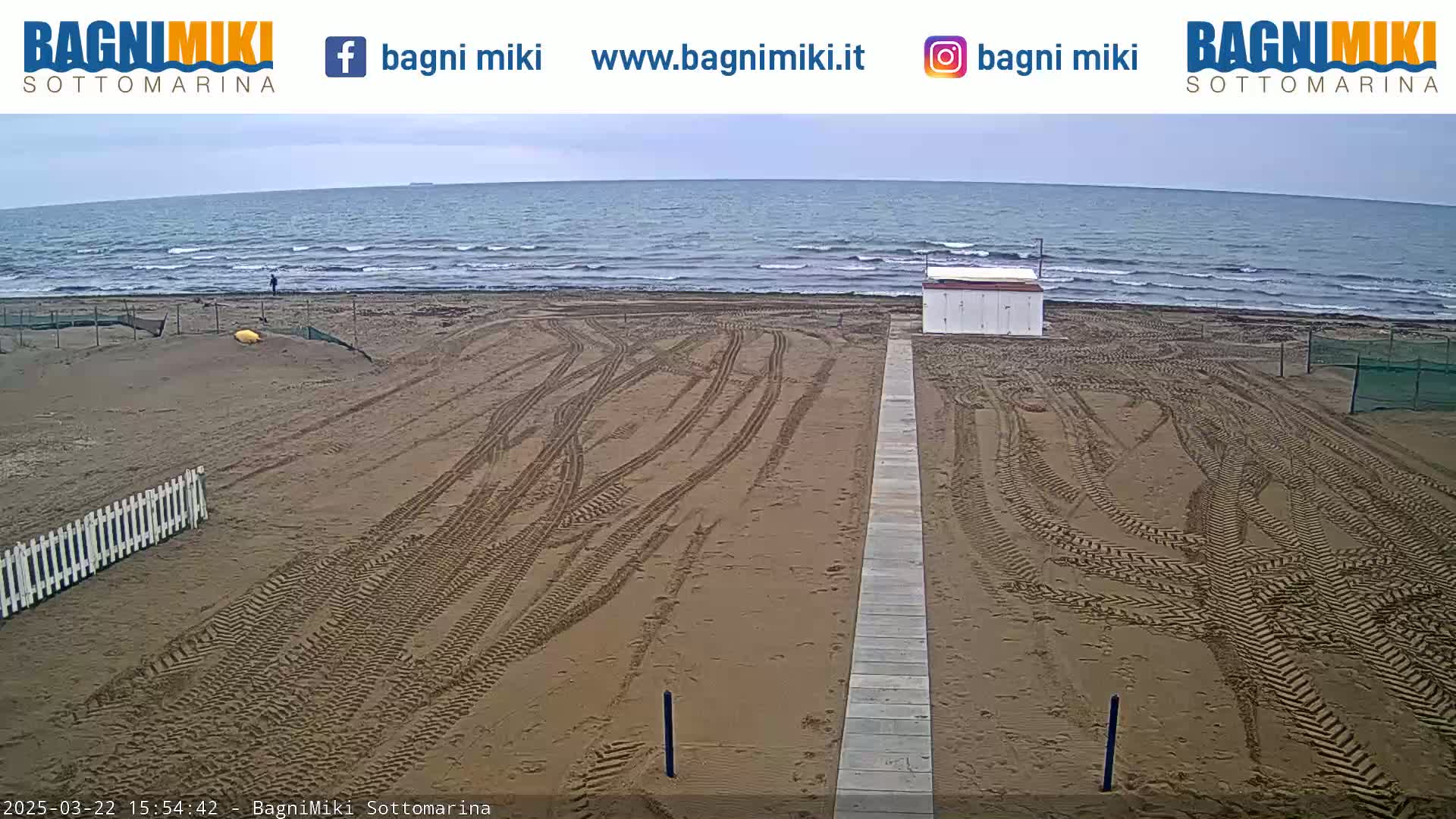 A sandy beach with tire tracks leading to a boardwalk and a small building, overlooking a wavy ocean under an overcast sky.