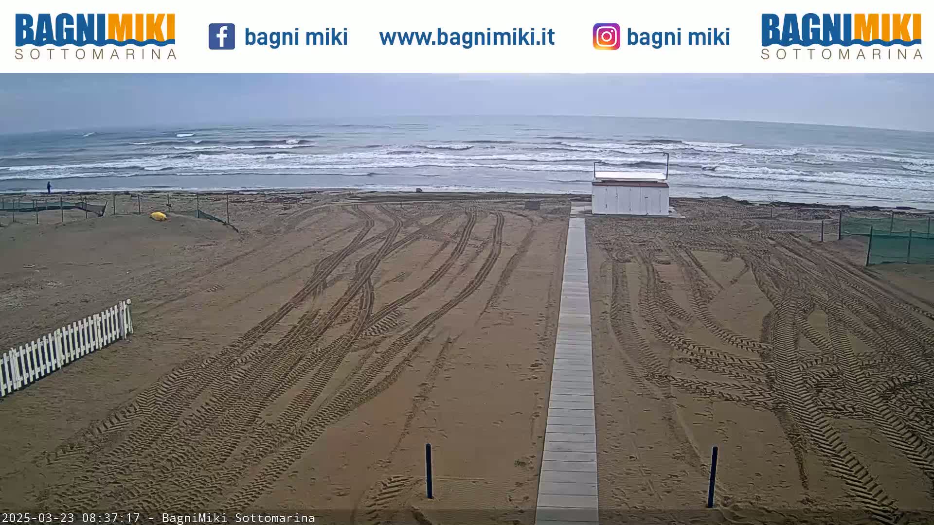 A sandy beach with tire tracks leading to a boardwalk, a small building, and an overcast sky over a somewhat choppy ocean.
