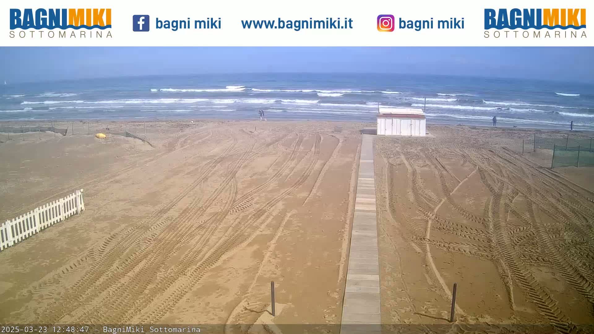 A sandy beach with tire tracks and a boardwalk leading to a small building is shown, with a wavy ocean under a clear sky in the background.