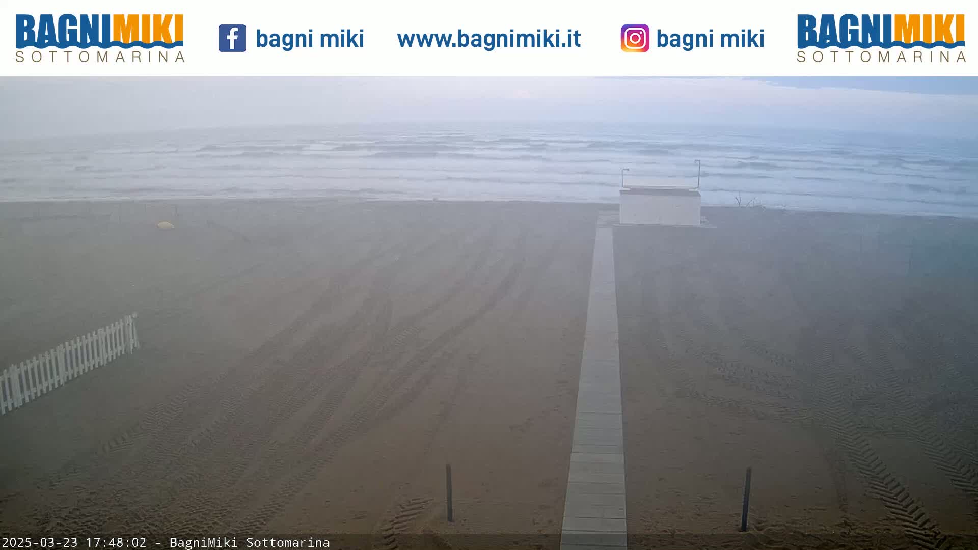A foggy beach scene shows a sandy shore with tire tracks, a walkway, and a small building, all under a hazy sky with a calm sea in the background.