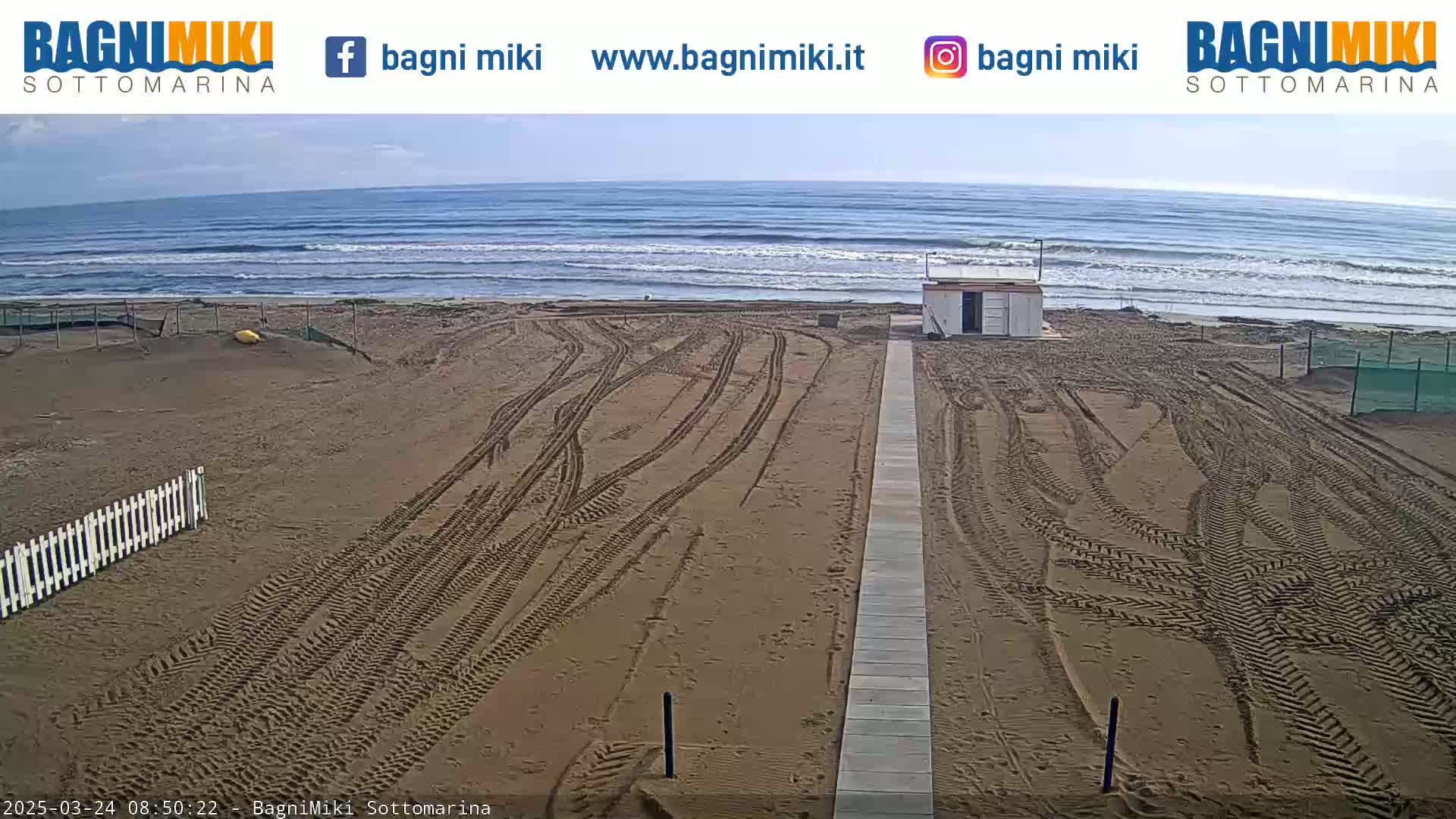 A sandy beach with tire tracks leading to a small building is visible, with a calm ocean in the background under a mostly clear sky.