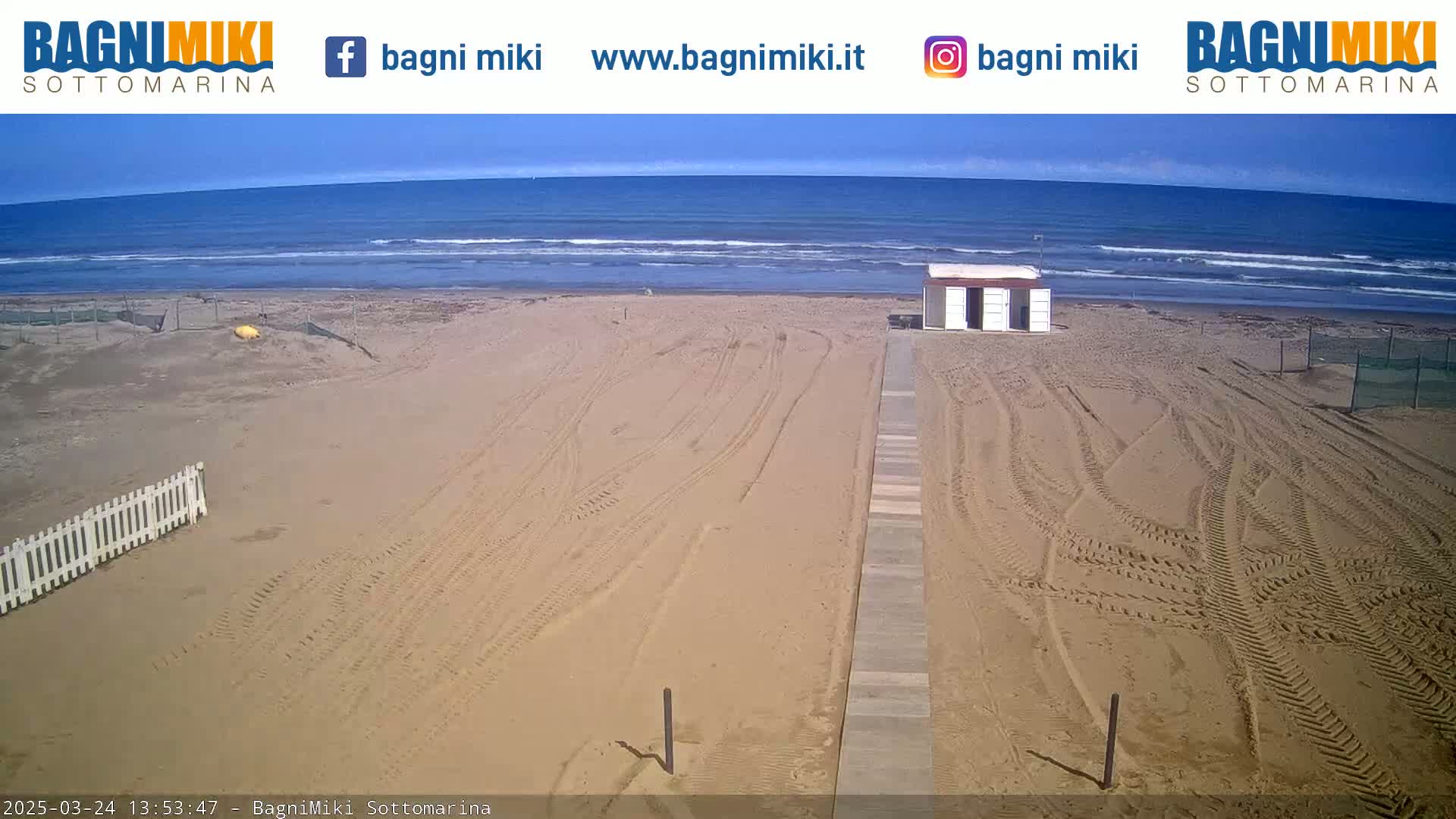 A sandy beach with tire tracks leading to a small building is shown on a mostly sunny day, with a calm ocean in the background.