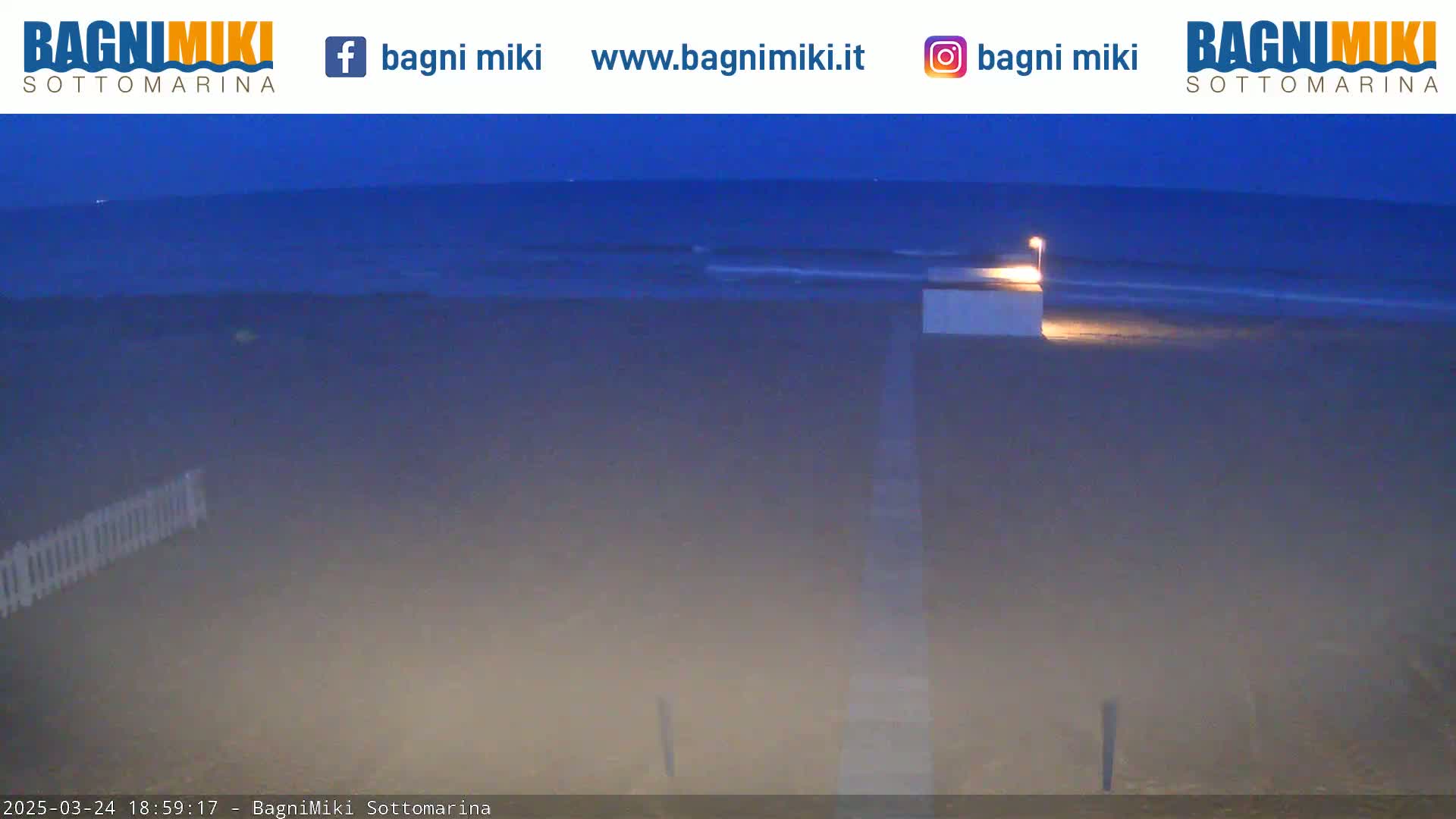 A dark beach at twilight shows a calm sea, a dimly lit structure on the sand, and a white fence in the foreground.