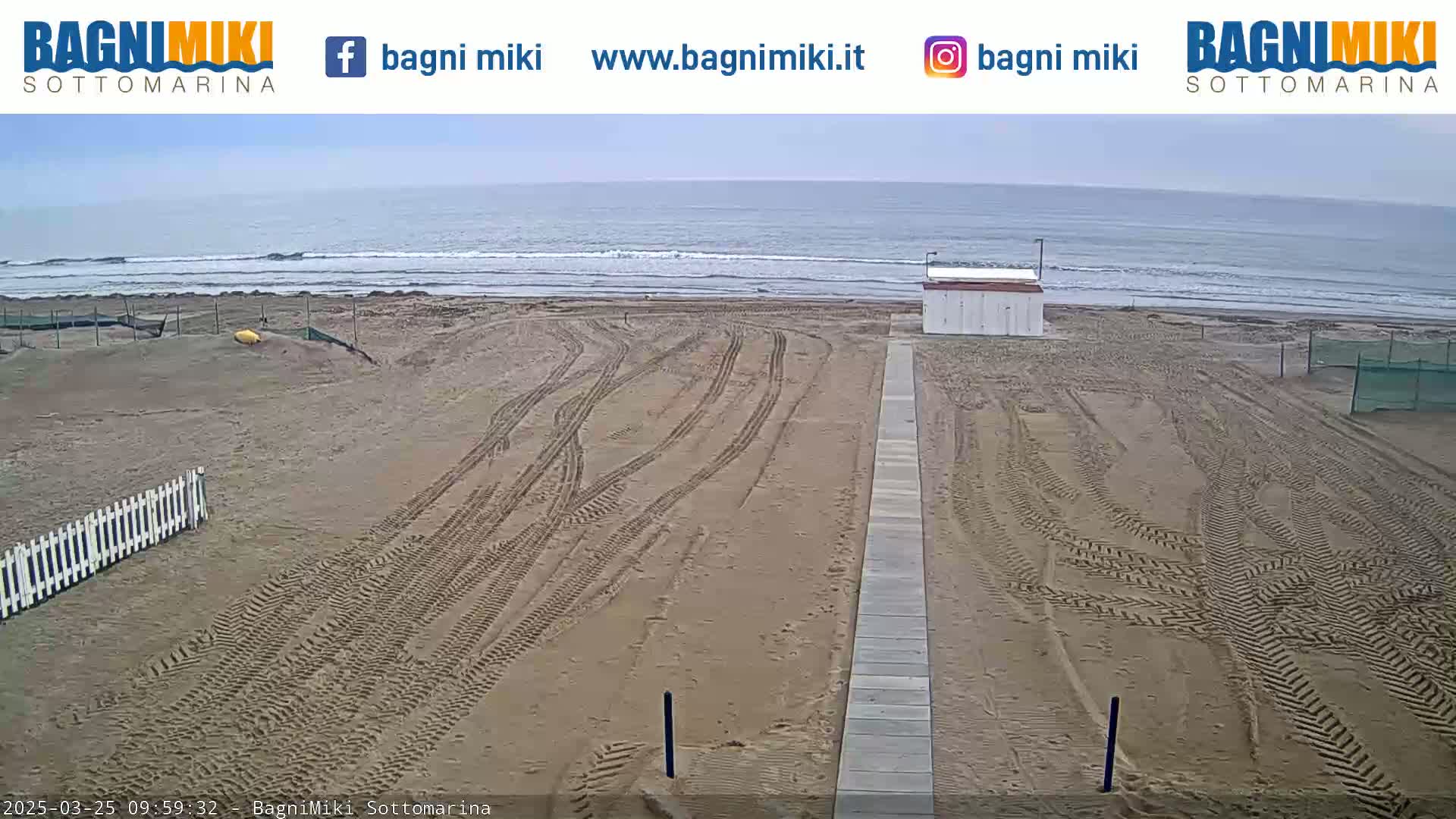 A mostly empty sandy beach with tire tracks and a small building is visible, overlooking a calm, gray ocean under an overcast sky.