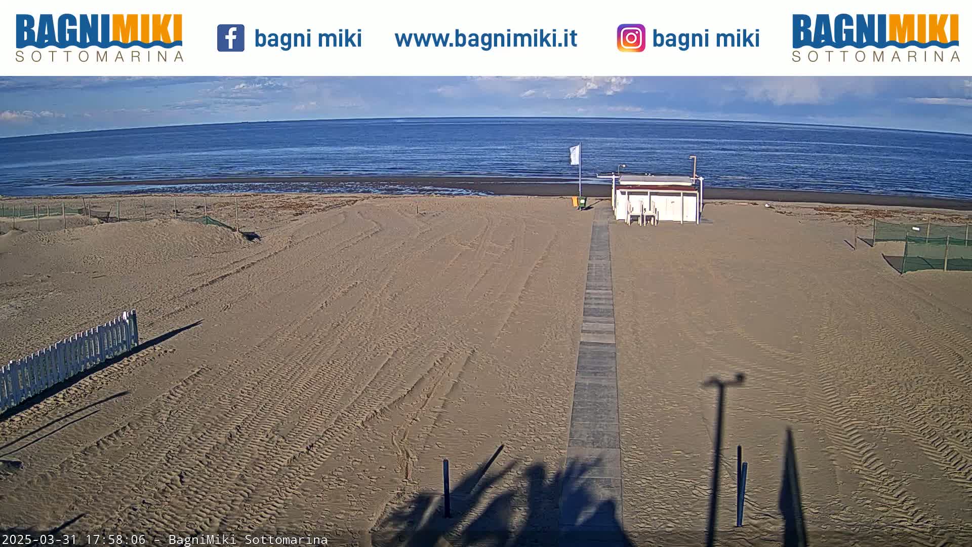 A sandy beach with a small building and a walkway leading to the water under a mostly clear sky.