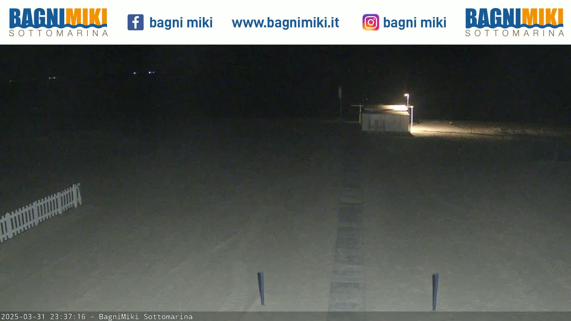 A dark, empty beach at night is illuminated by a single light near a small building.