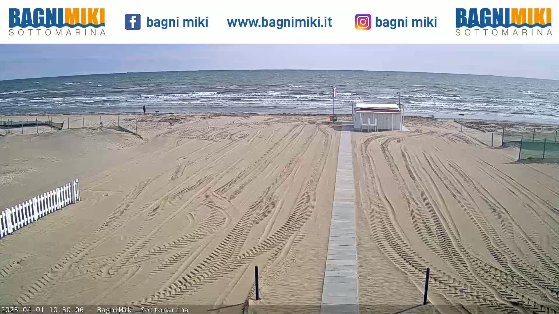 A sandy beach with tire tracks leading to a small white building is shown, with a somewhat wavy ocean under a partly sunny sky.