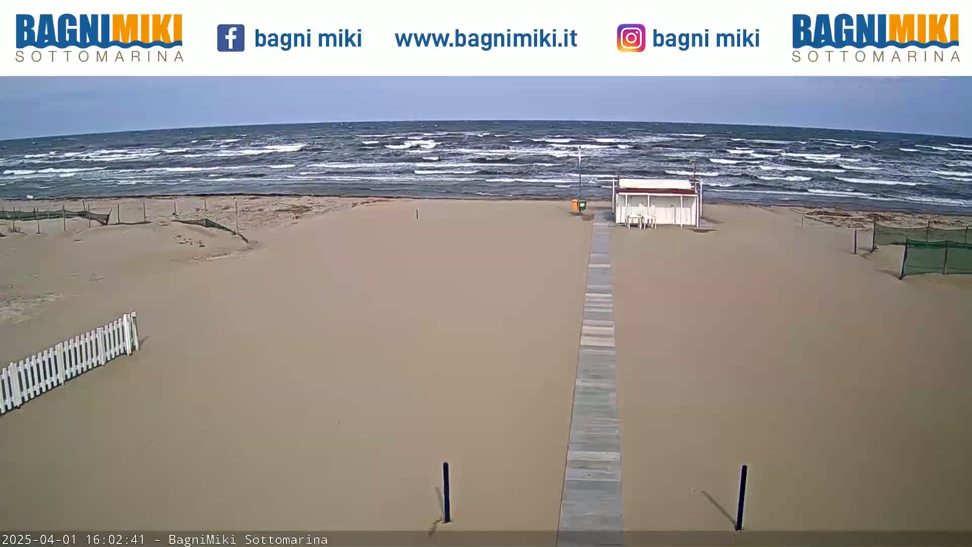 A sandy beach with a boardwalk leading to a small building is visible, with a somewhat choppy sea under a mostly clear sky.