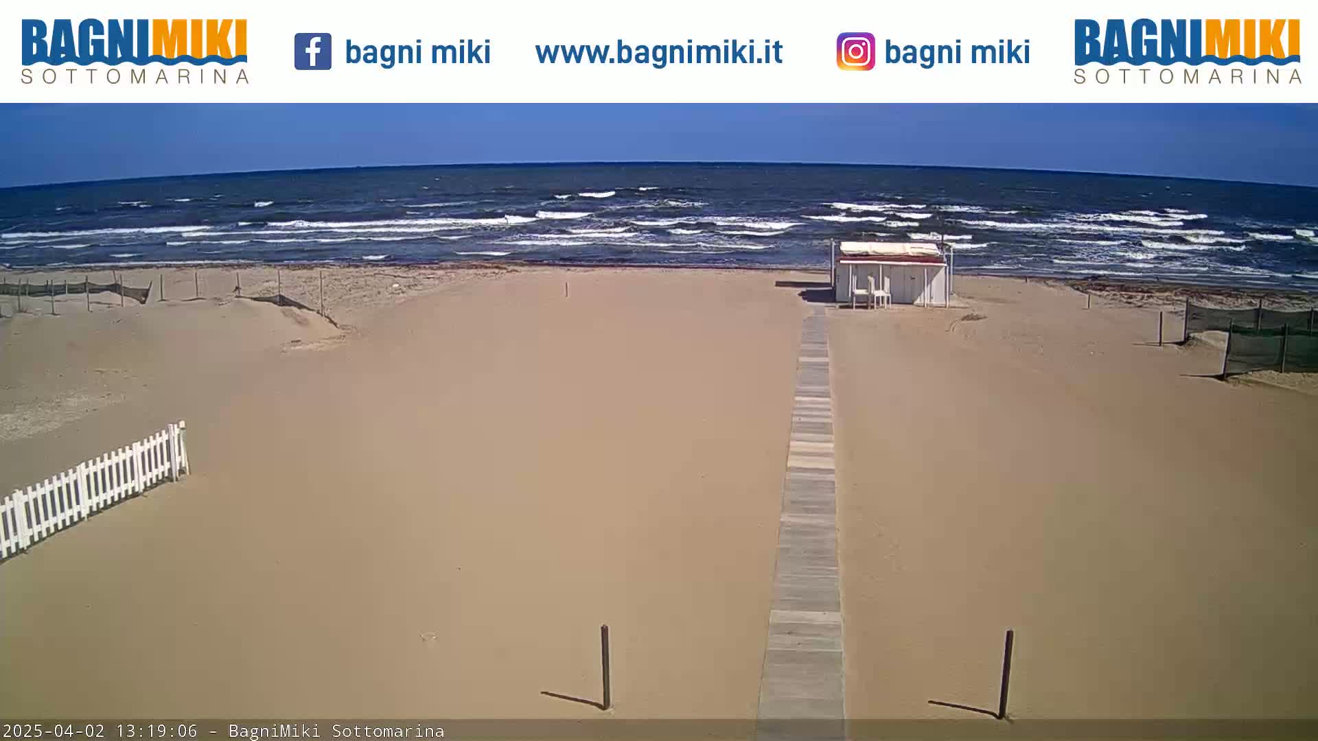 A sandy beach with a boardwalk leading to a small structure is visible, with a somewhat choppy ocean under a sunny sky in the background.