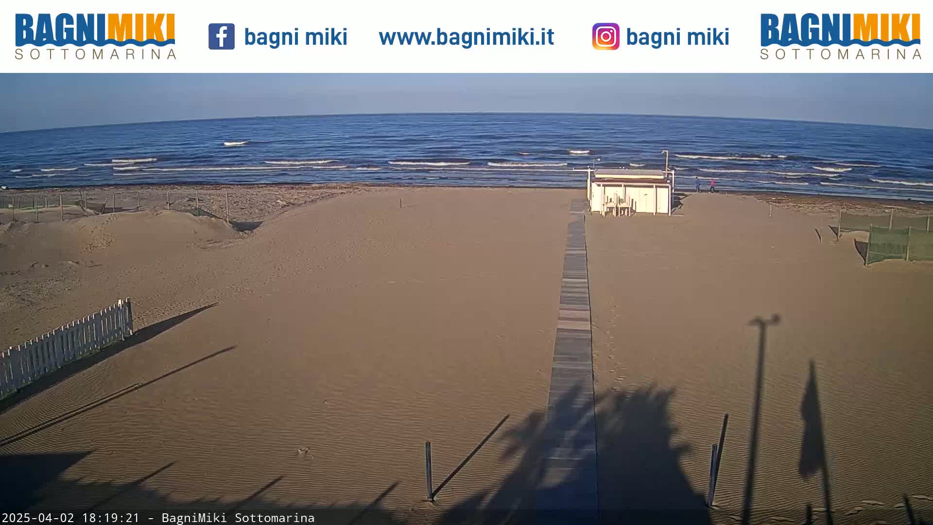 A sandy beach with a boardwalk leading to a small building, under a clear sky, and a calm ocean with small waves in the background.