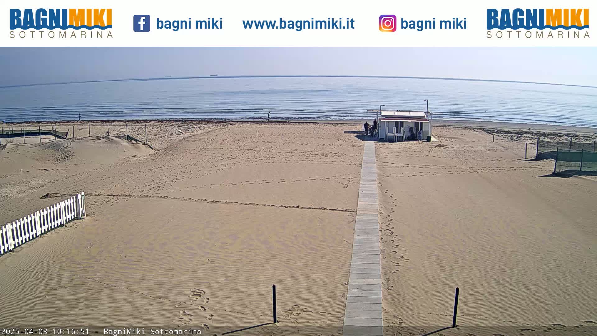 A mostly empty sandy beach with a small building and a boardwalk stretches toward a calm, sunny ocean.