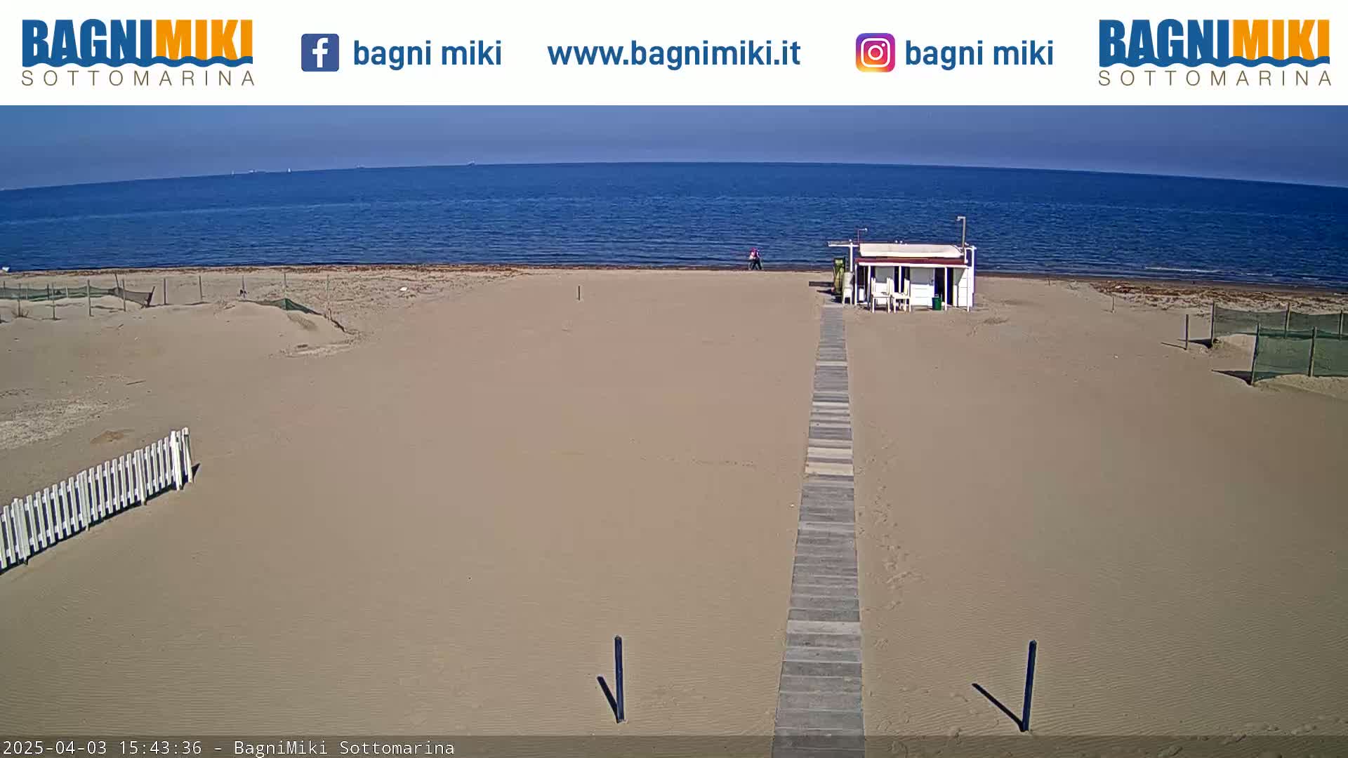 A sandy beach with a boardwalk leading to a small building, under a clear sunny sky and calm ocean.