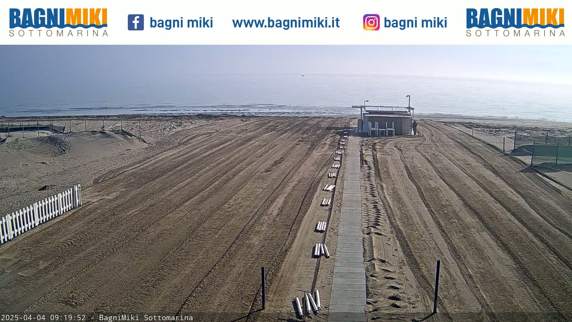 A sandy beach with a paved walkway leading to a small building, under a mostly clear sky.
