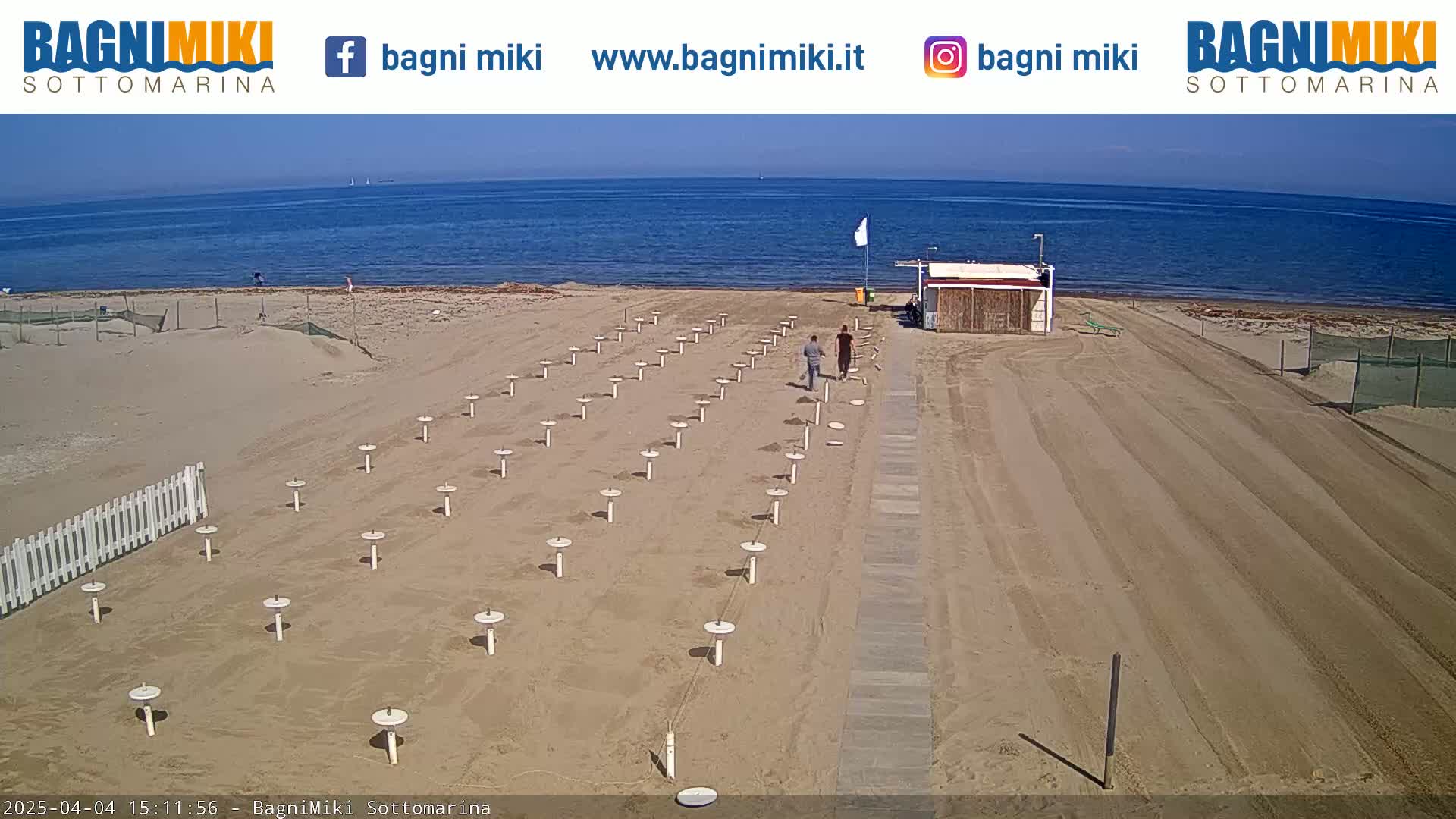 A sandy beach with rows of umbrella stands, a small building, and two people walking near a calm, blue ocean under a sunny sky.