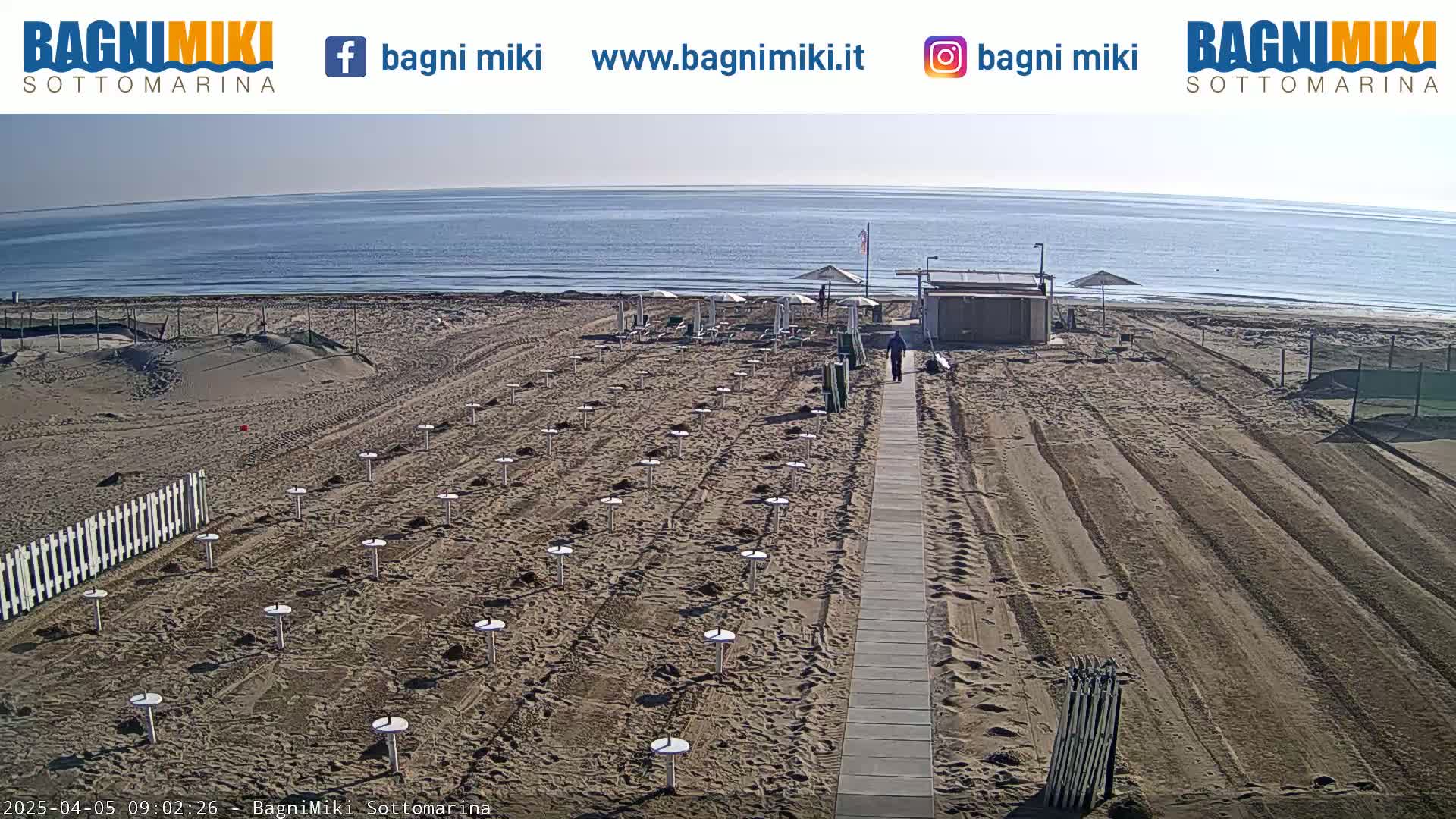 A sandy beach with a boardwalk, beach umbrellas set up in rows, a small building, and calm ocean under a clear sky.