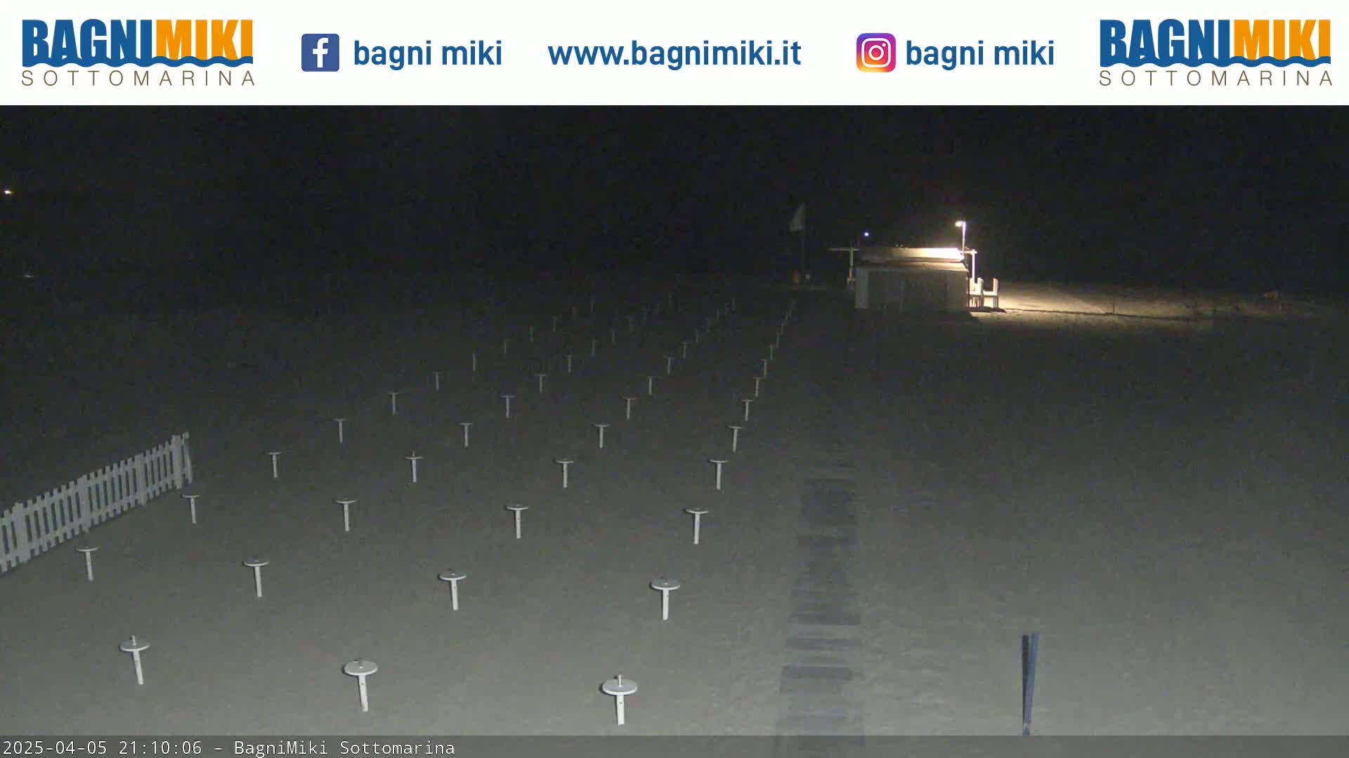 At night, a sandy beach is sparsely populated with numerous short posts and a white fence, illuminated by a single lamppost.