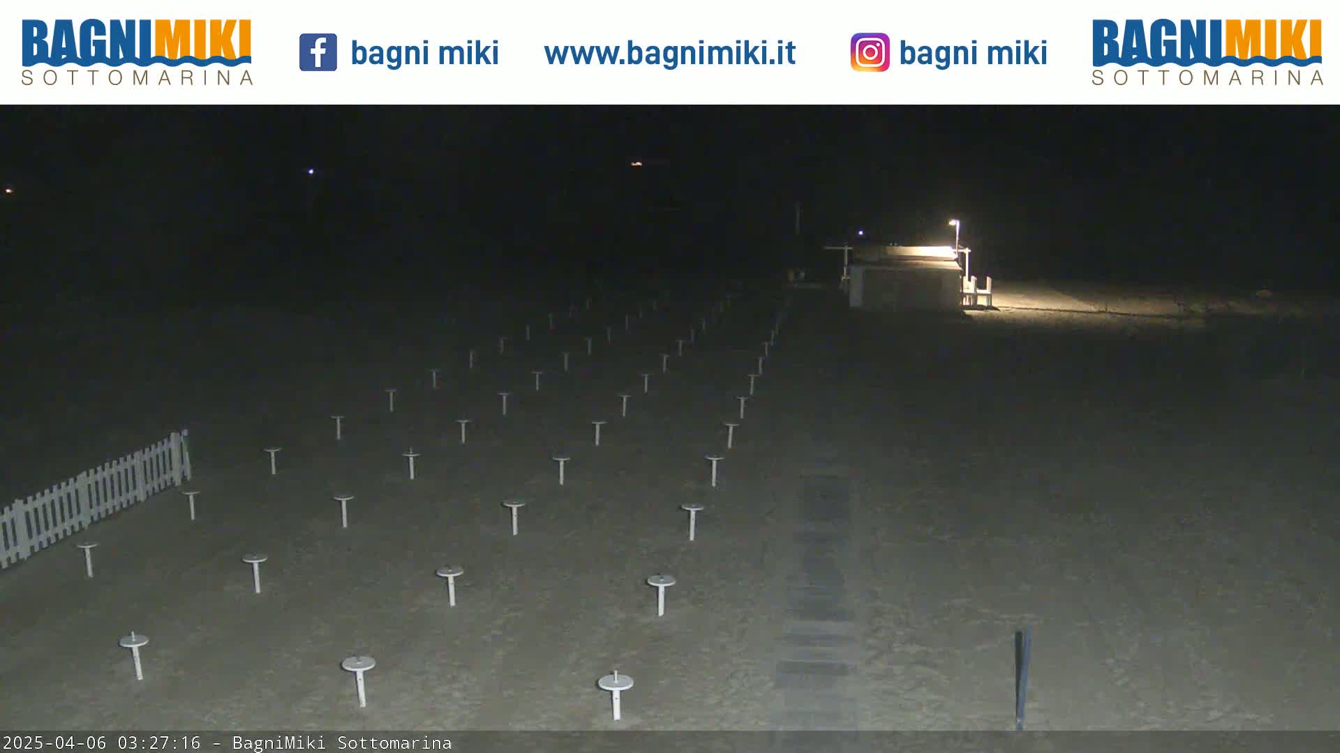 Numerous empty beach umbrella stands are arranged on a dark, sandy beach at night, illuminated by a single lamppost.