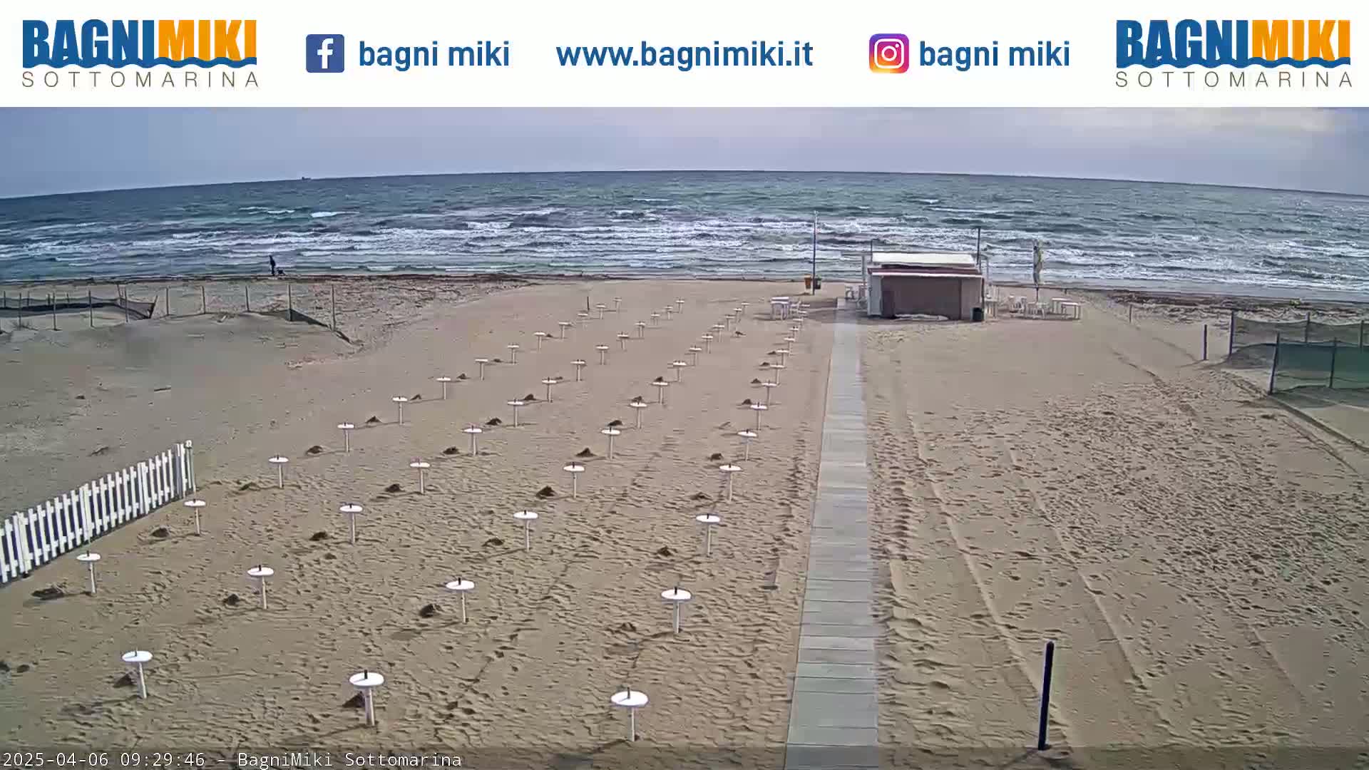 A sandy beach with numerous empty umbrella stands is shown, with a somewhat choppy ocean in the background under a mostly clear sky.