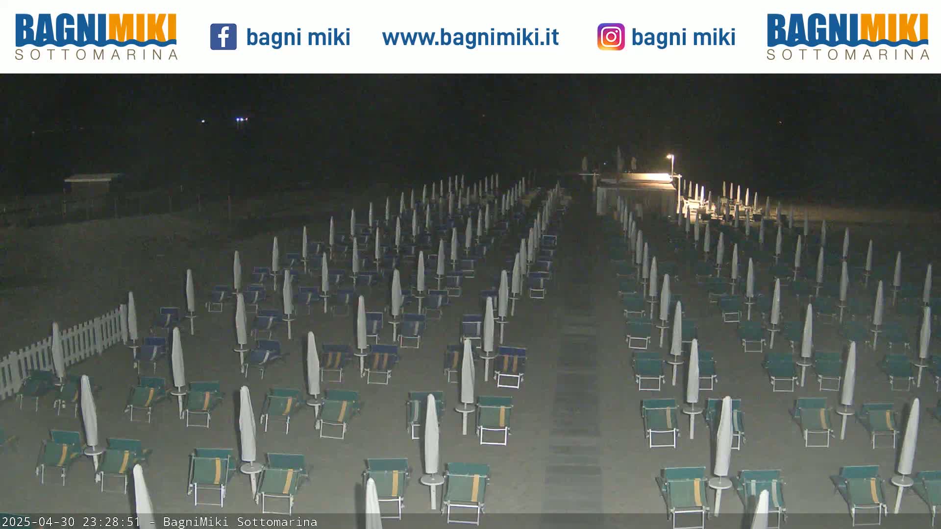 A nighttime beach scene shows numerous closed beach umbrellas and lounge chairs arranged in neat rows on a sandy beach.