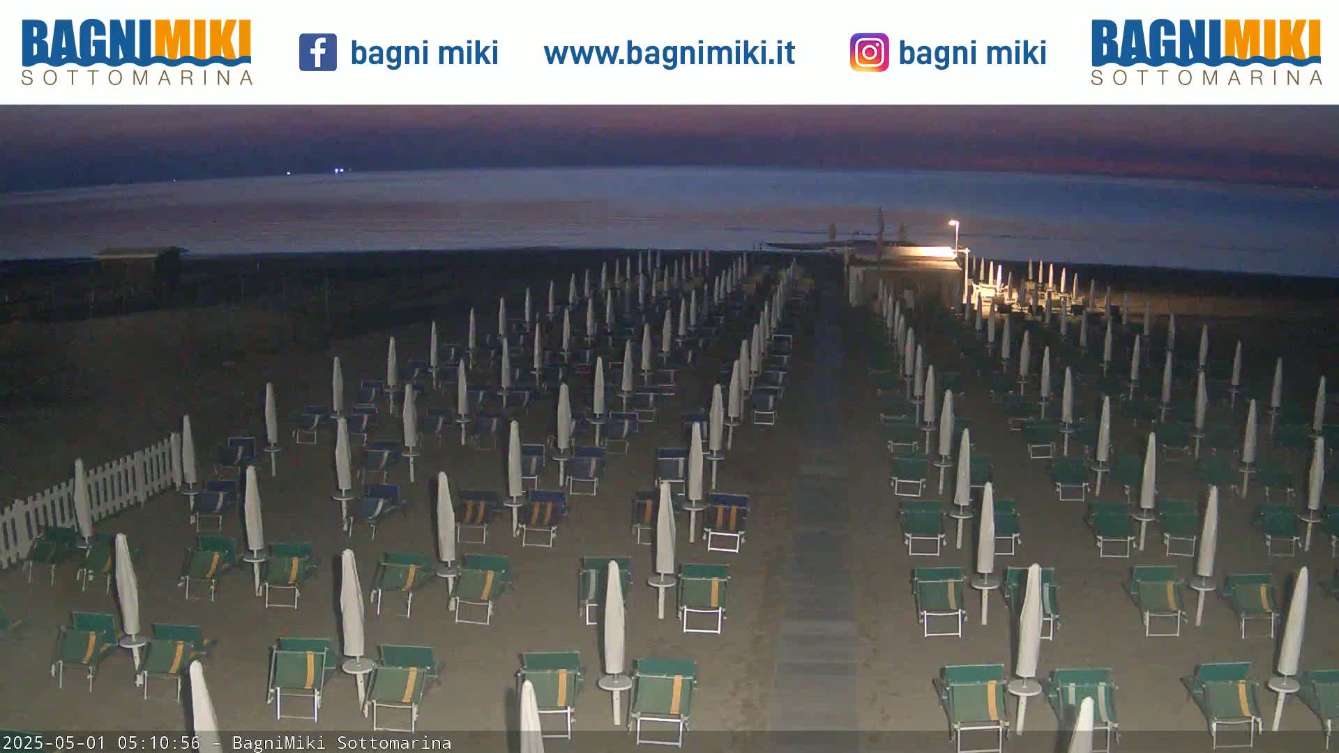 Rows of beach umbrellas and lounge chairs stand on a dark, sandy beach at dusk, overlooking a calm sea under a twilight sky.