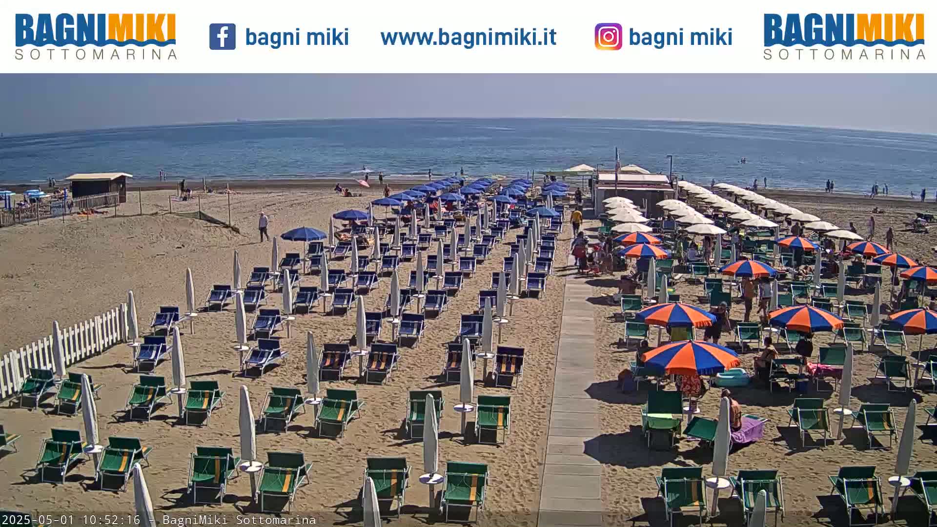 A sandy beach with numerous beach umbrellas and lounge chairs under a sunny sky, next to a calm ocean.