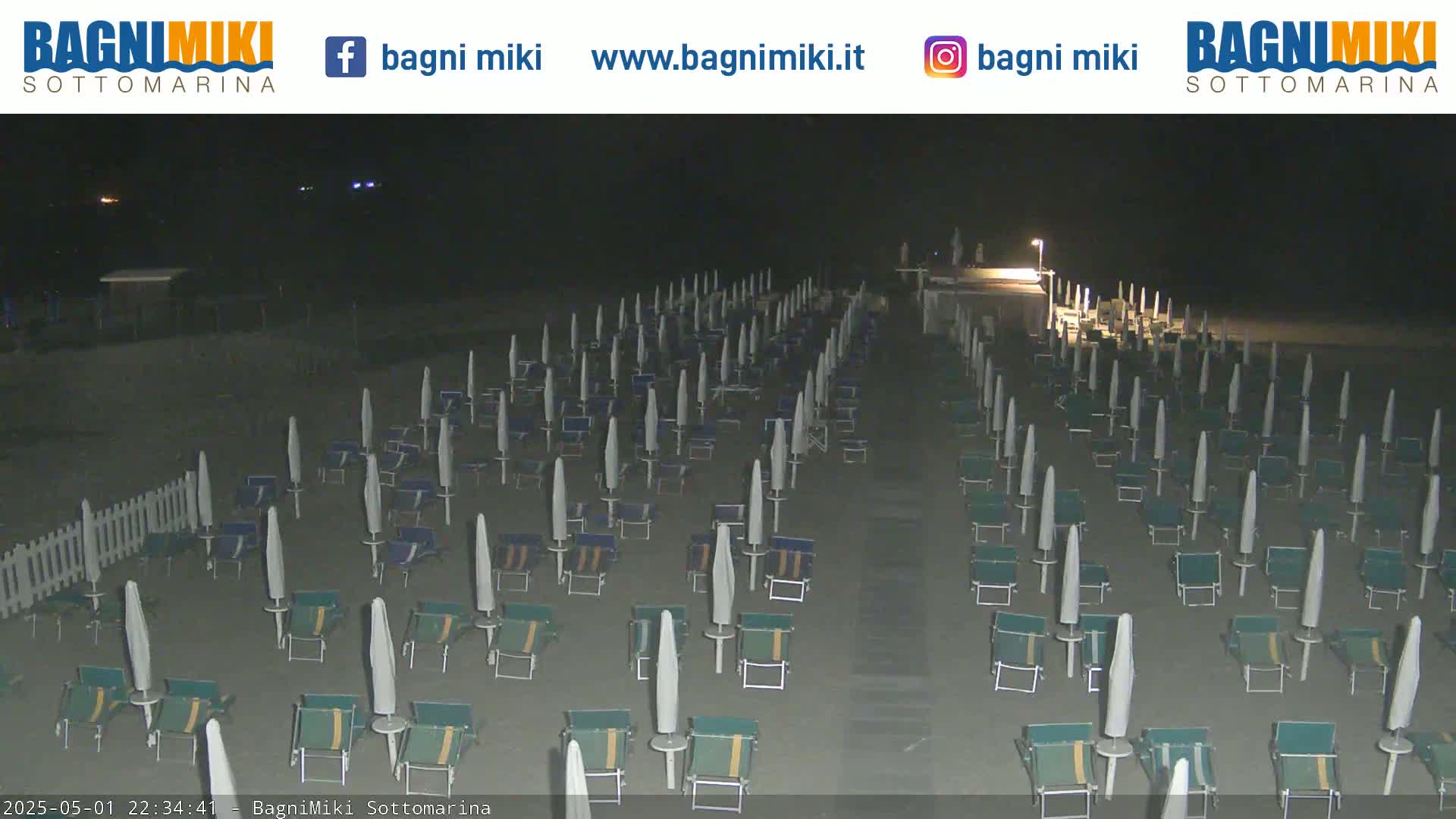 Rows of closed beach umbrellas and lounge chairs sit on a dark sandy beach at night.