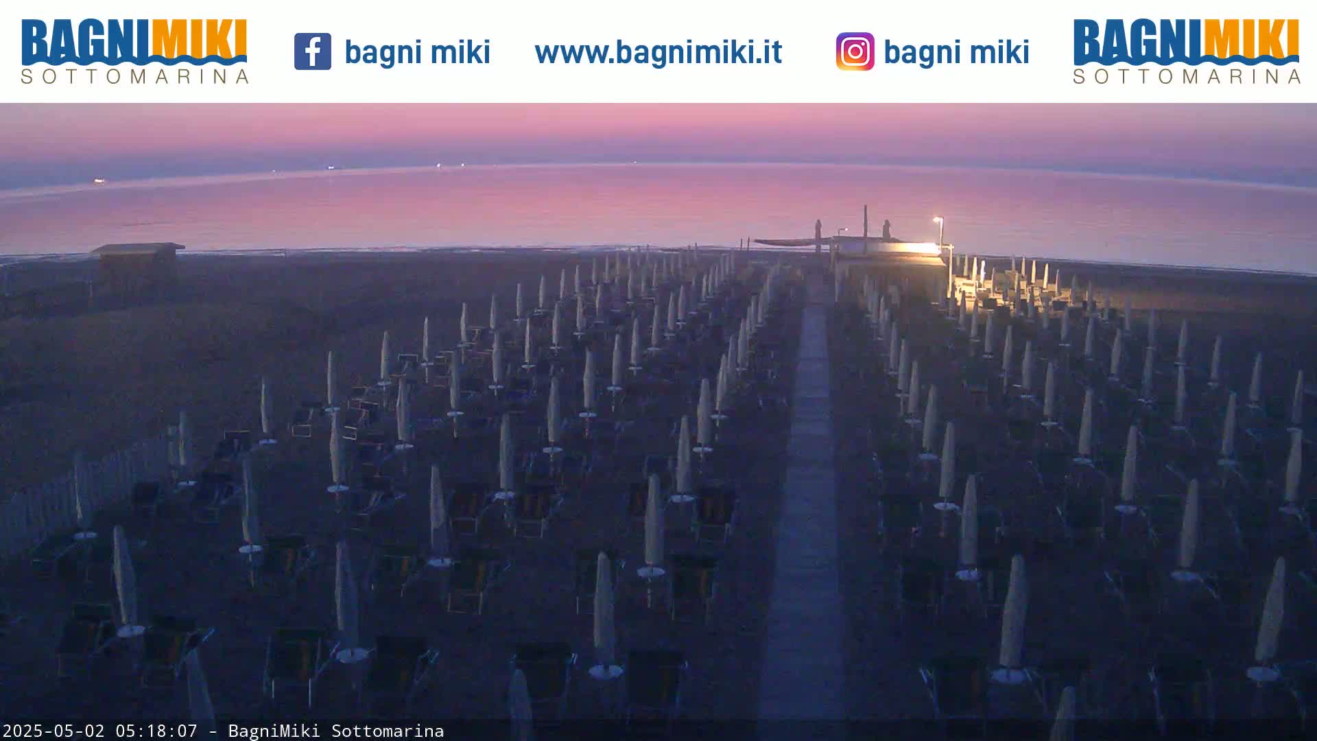 Rows of beach umbrellas and lounge chairs sit on a dark beach at dusk under a pink and purple sky.