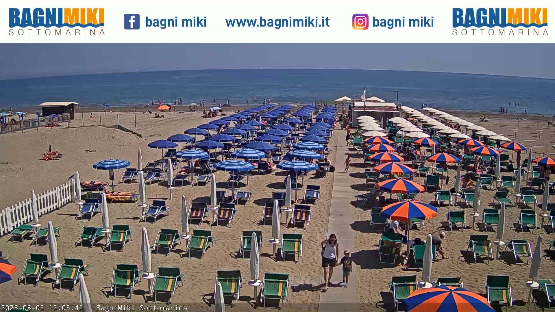 A sandy beach with numerous blue and orange umbrellas and lounge chairs lines a calm ocean under a sunny sky.